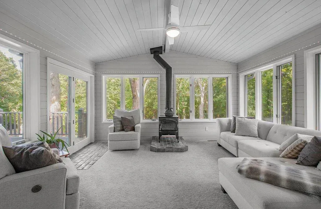 Bright sunroom with large windows, gray seating, and a central black wood stove.
