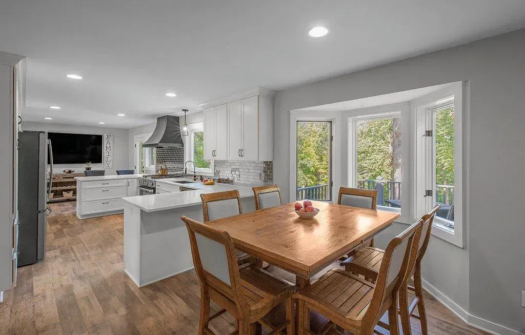 Bright dining area with wooden table, chairs, kitchen island, and large windows overlooking greenery.