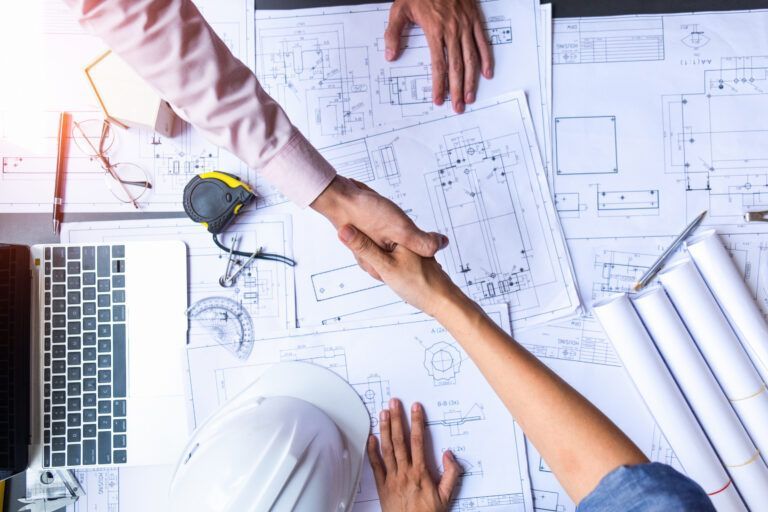 Colleagues shaking hands over architectural blueprints beside a laptop and measuring tools