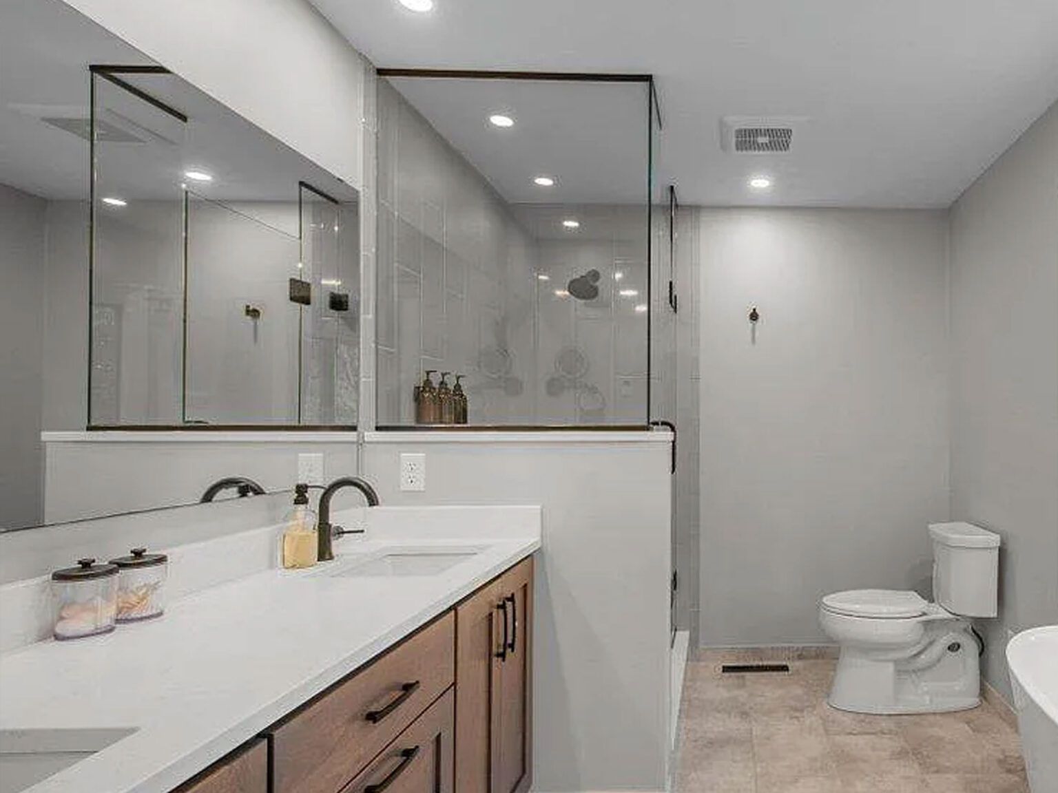 Modern bathroom with white vanity, large mirror, glass shower, and toilet on a light tile floor