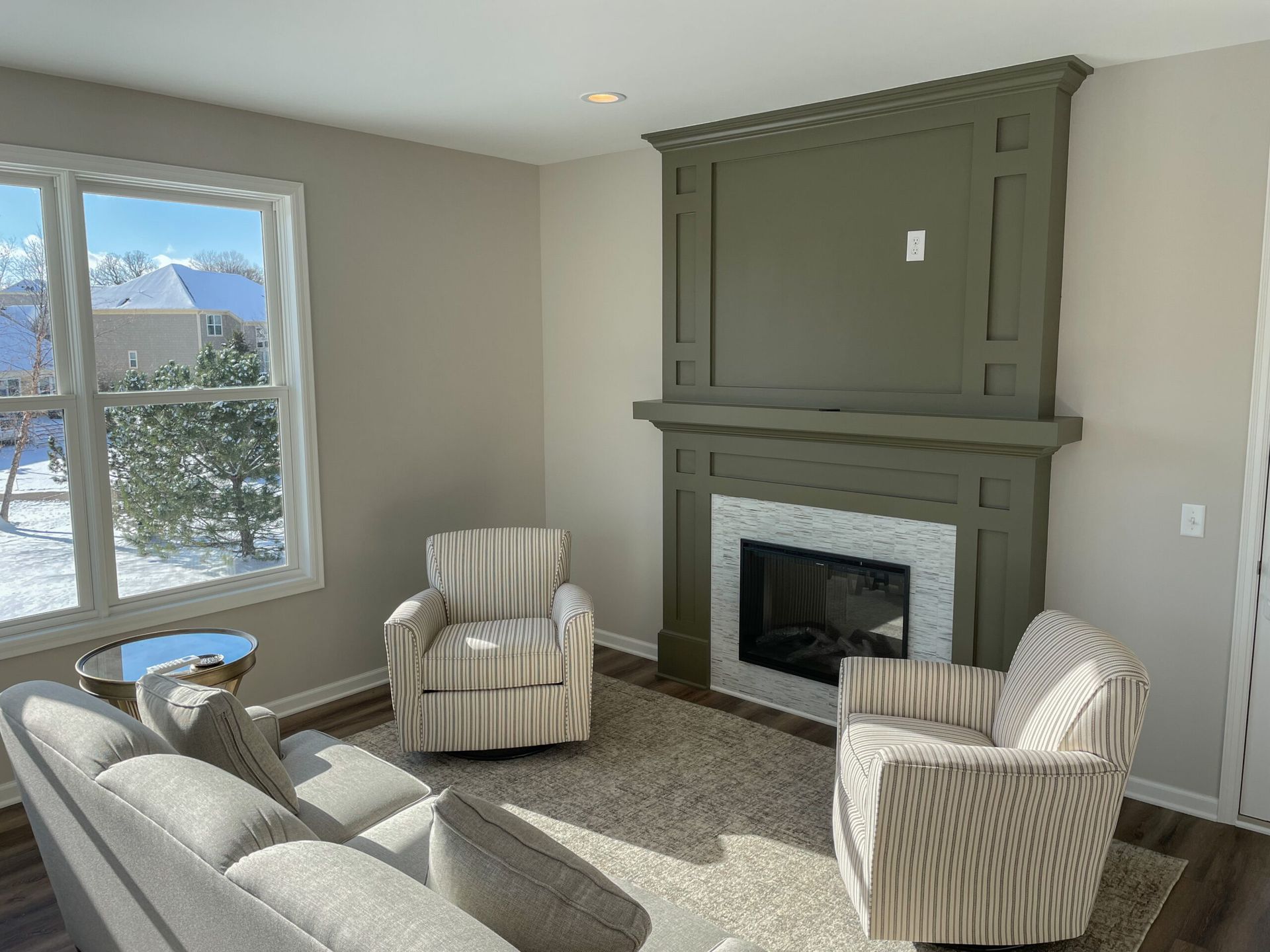 Bright living room with sage-green fireplace, striped chairs, and a large window overlooking snow outside