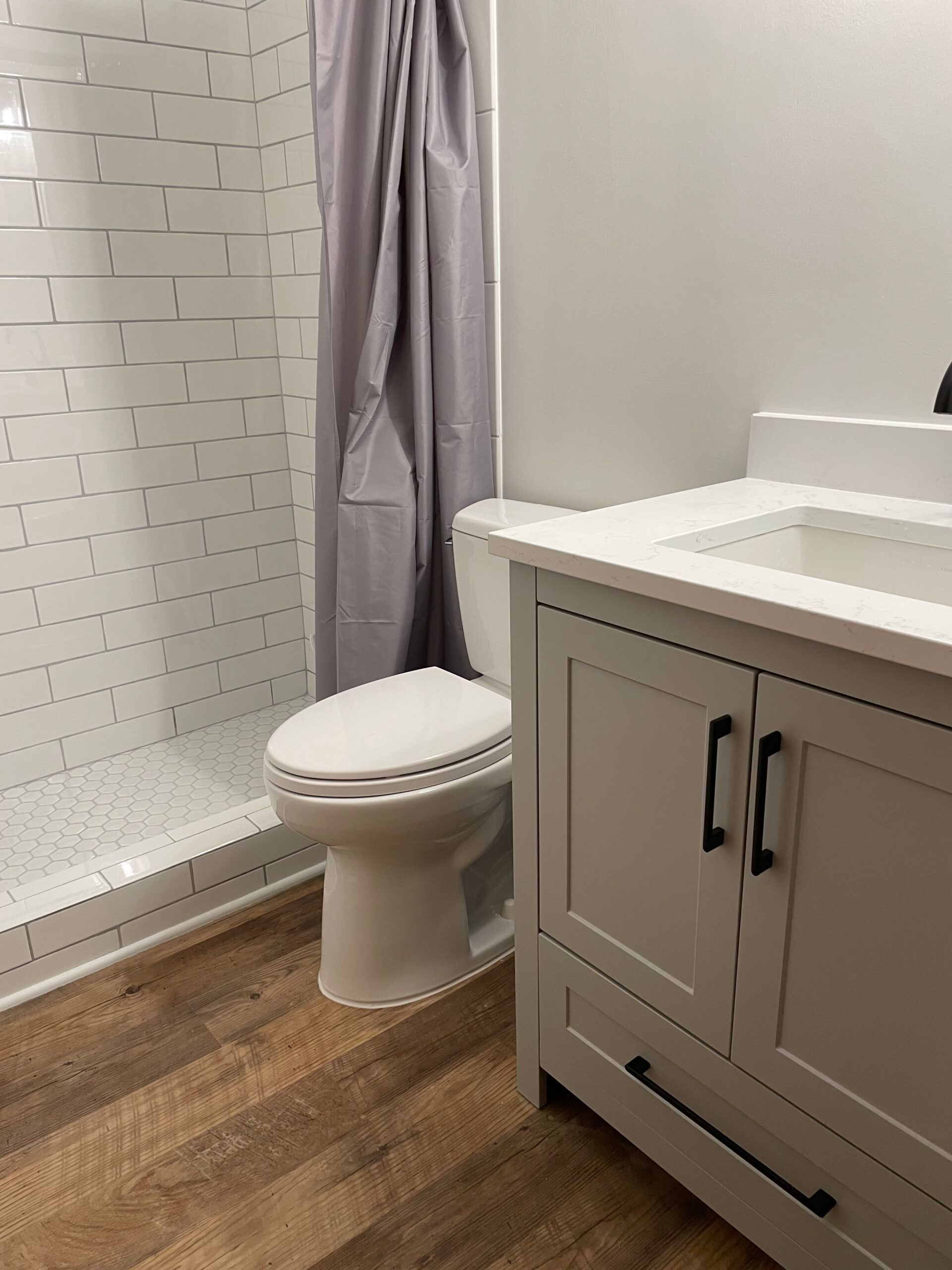 Bathroom with white toilet, shower, and vanity on wood floor, with a gray curtain.