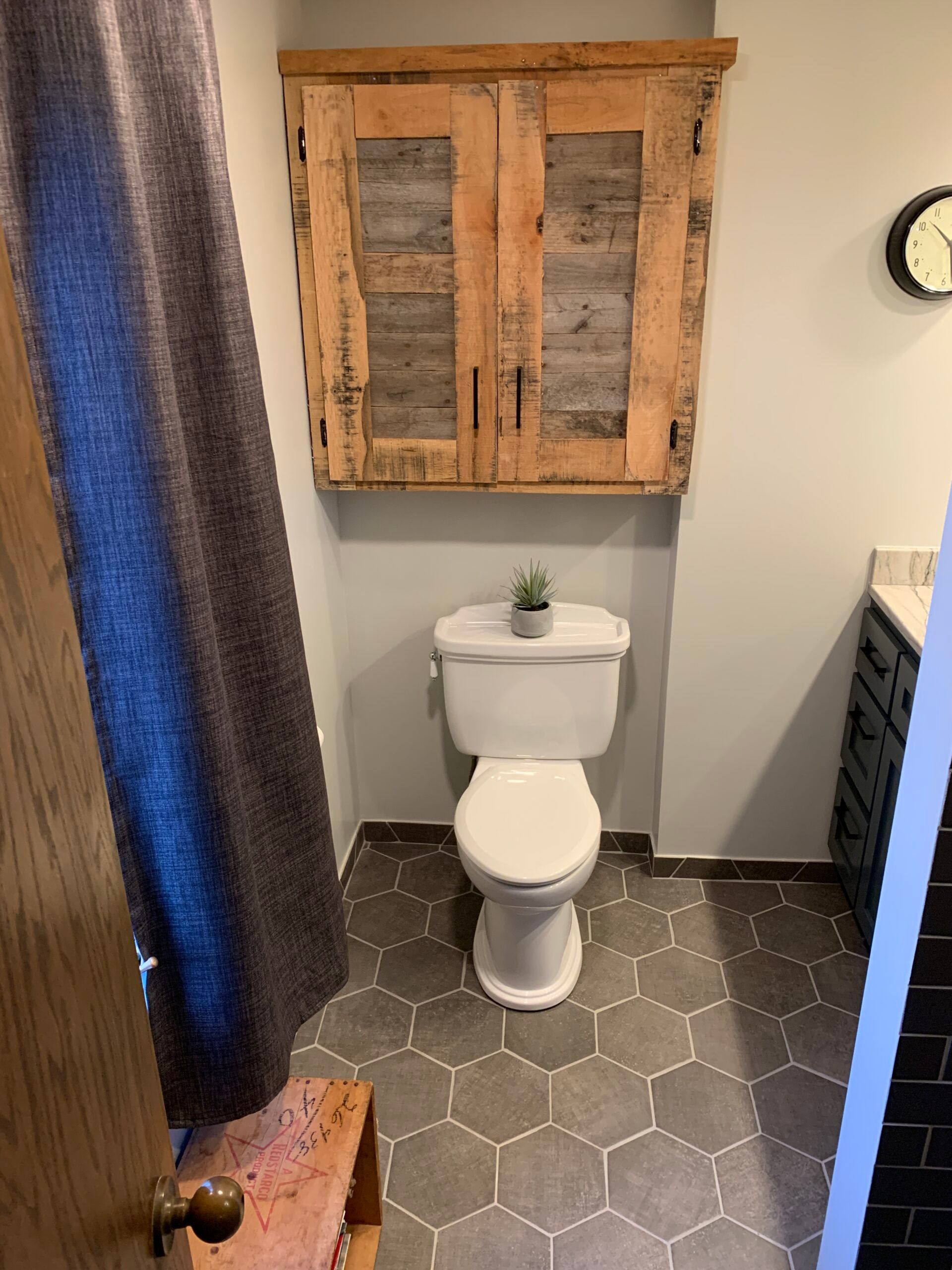 Small bathroom with a toilet, rustic wall cabinet, gray hex tile floor, and shower curtain.