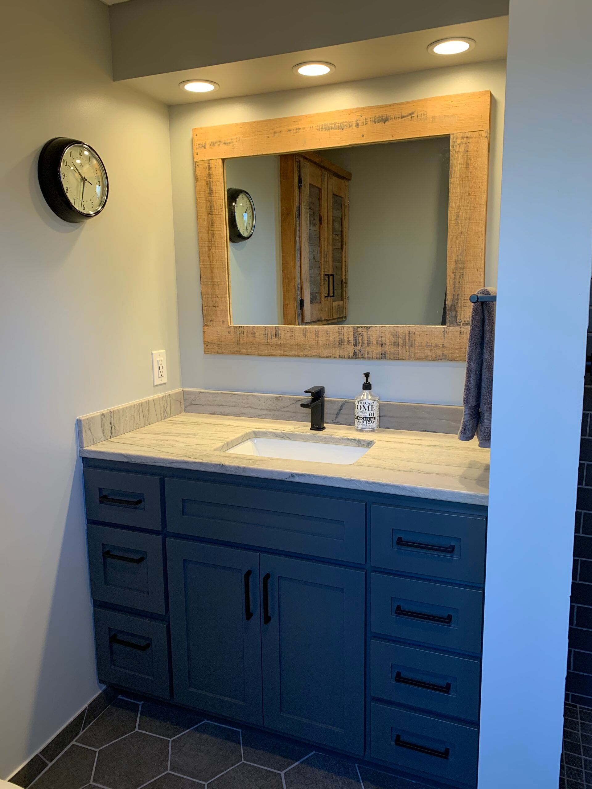 Blue bathroom vanity with marble countertop, round mirror, wall clock, and recessed lighting
