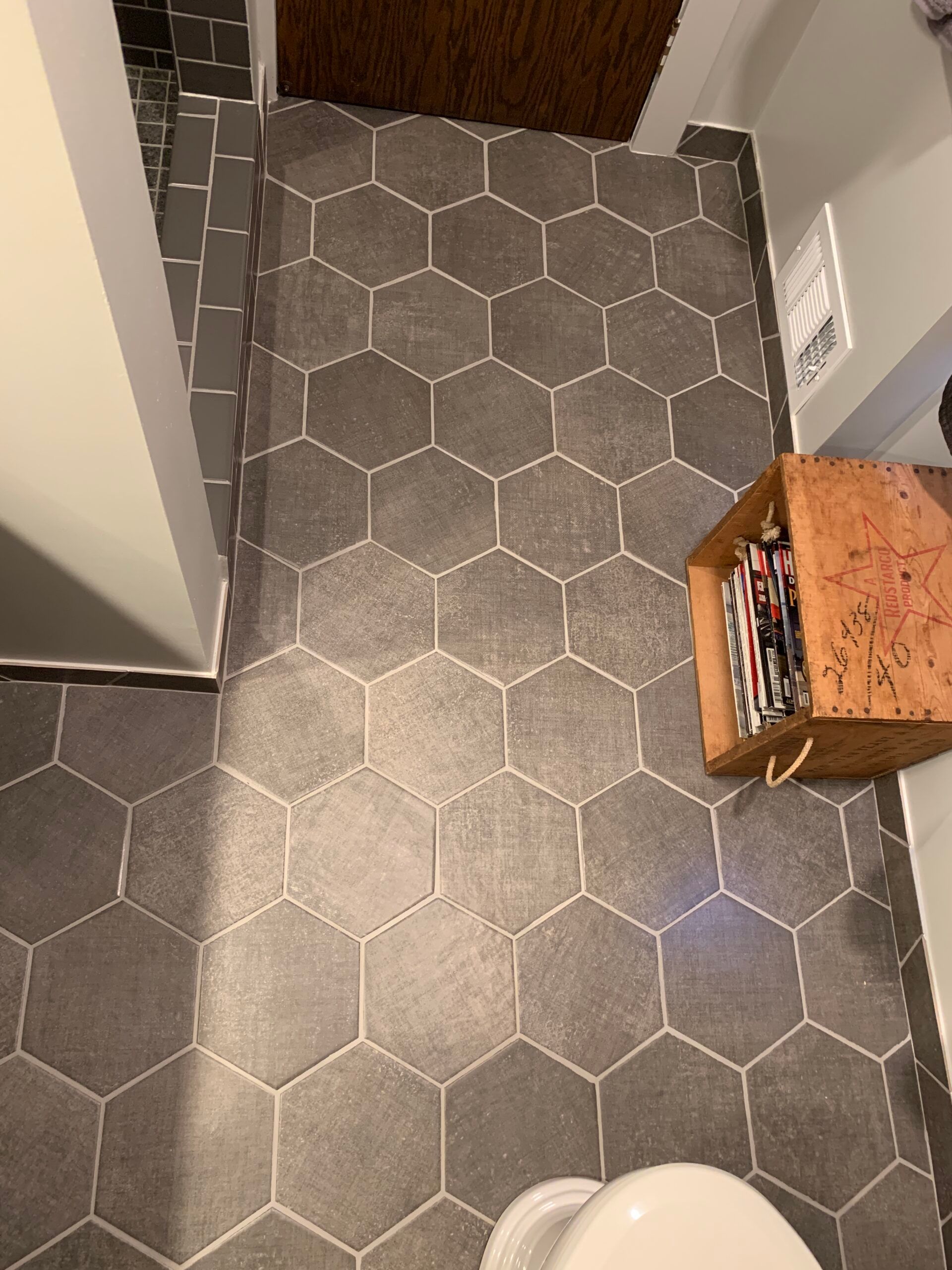 Narrow hallway with dark hex tile floor, a wooden crate on the right, and a brown rug at the far end.