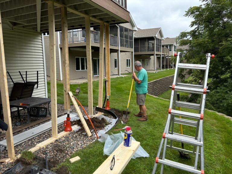 Person shoveling dirt beside a house porch with tools, ladder, and gravel in the yard