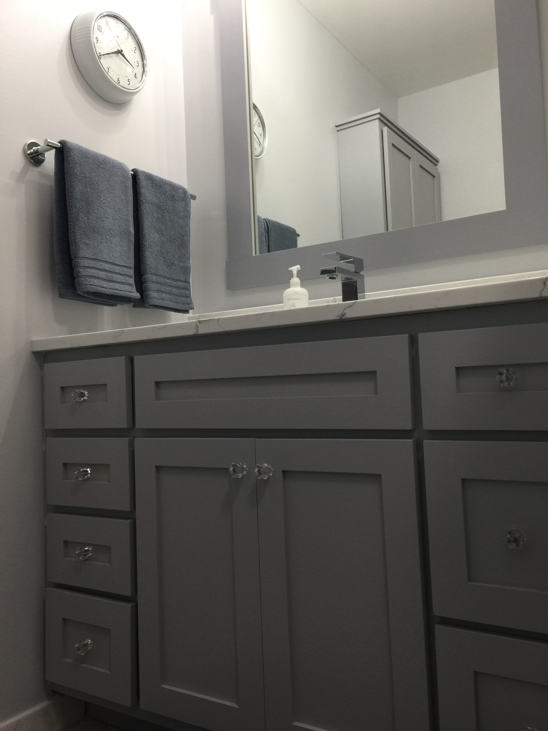 Gray bathroom vanity with mirror, sink, towel rack, and wall clock above.