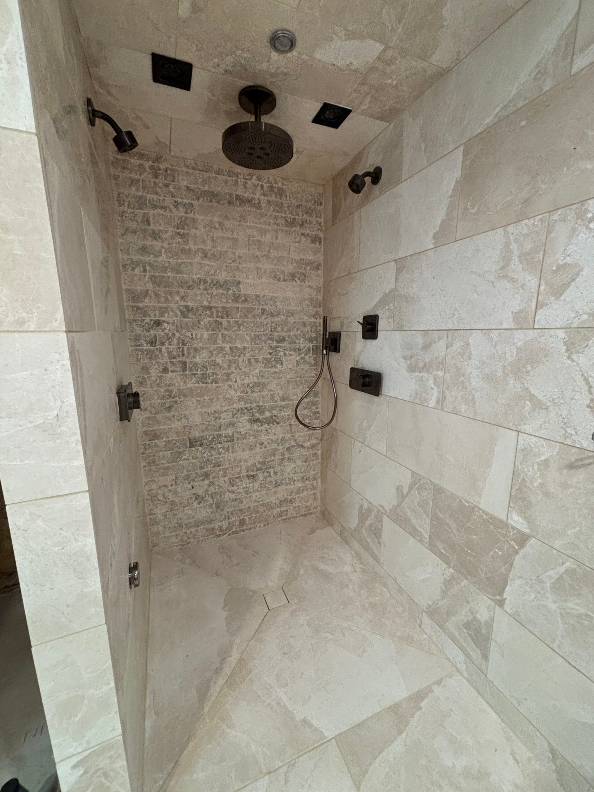 Empty tiled shower with multiple black showerheads and a brick accent wall.