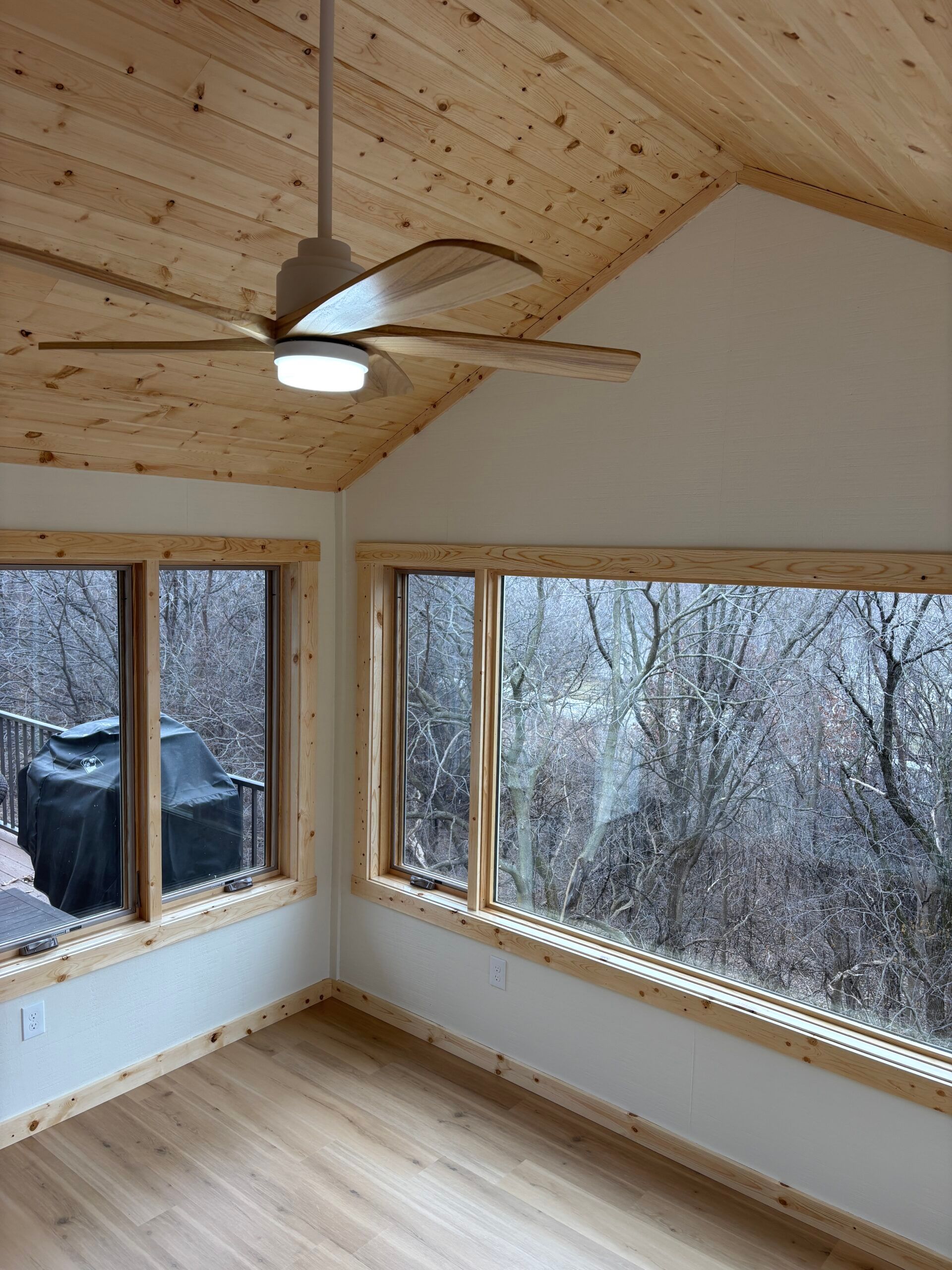 Cozy room with wood ceiling, ceiling fan, large windows, and snowy trees outside.