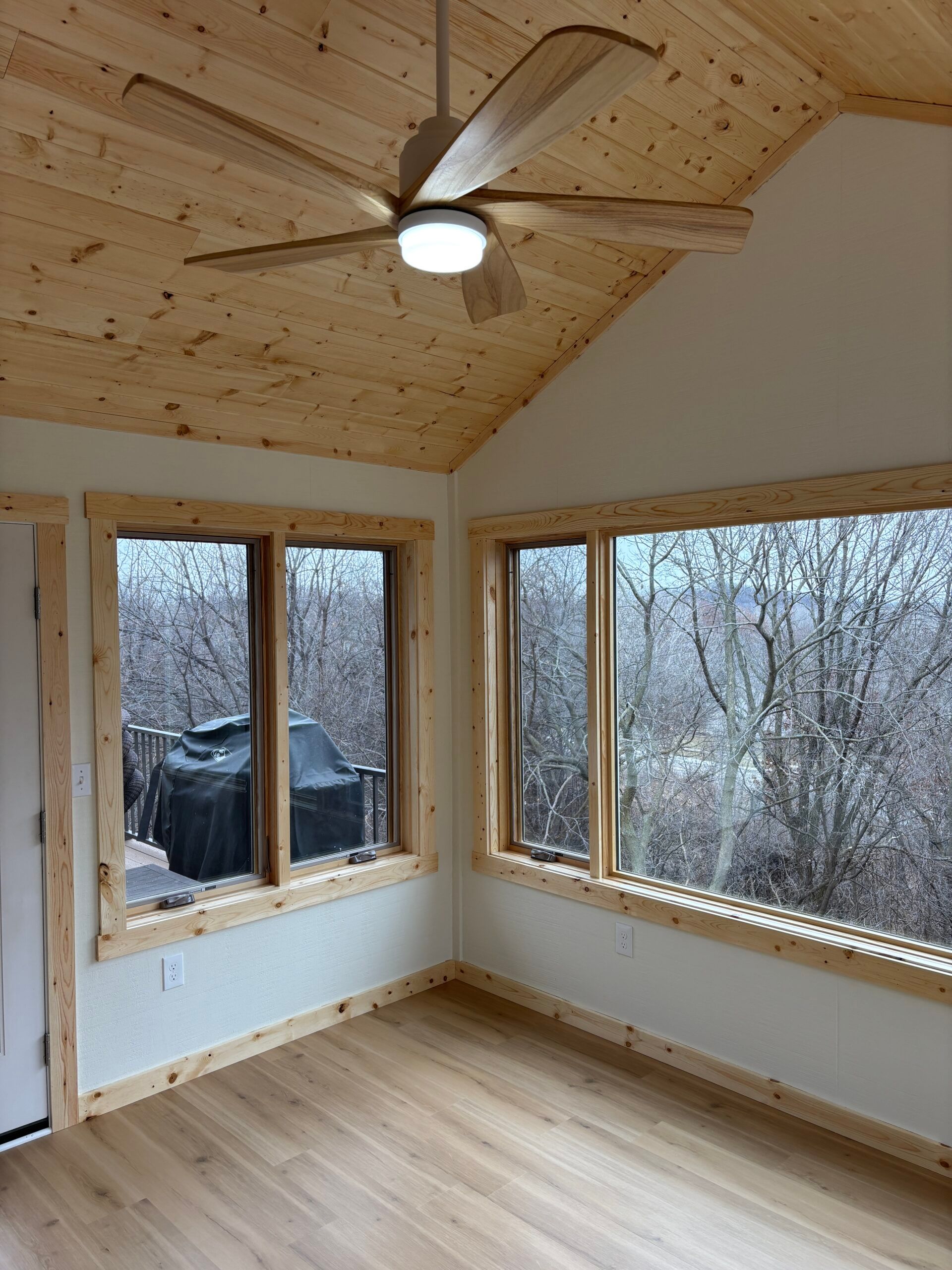 Empty sunroom with wood ceiling, light wood floor, and large corner windows overlooking bare trees.