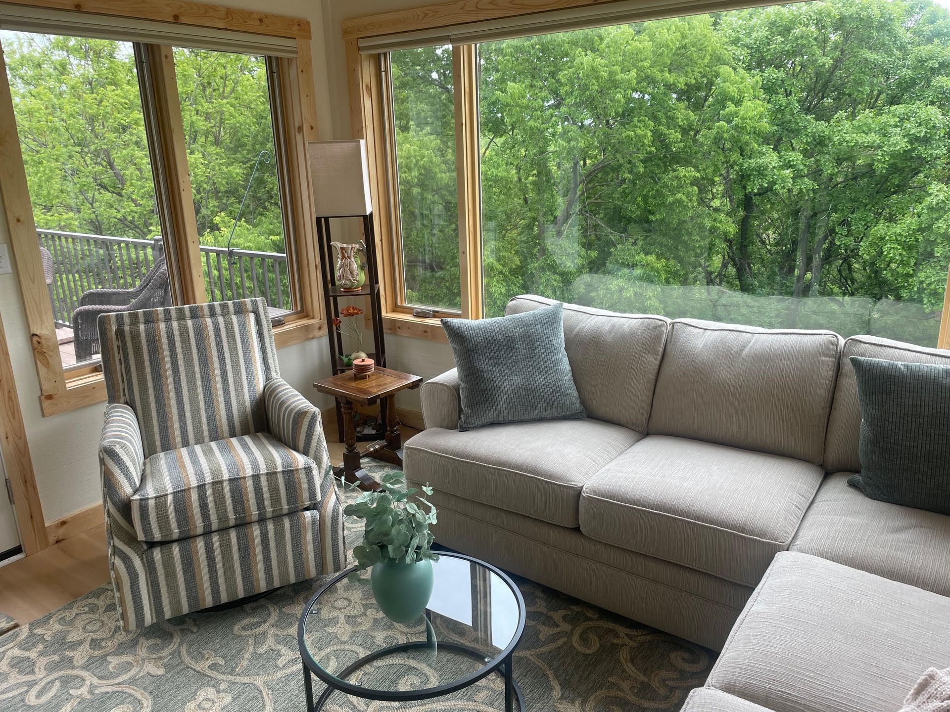 Bright sunroom with a gray sectional, striped armchair, glass coffee table, and large windows overlooking trees