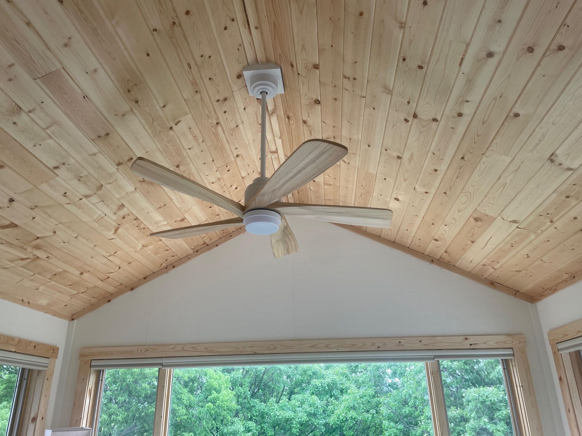 Ceiling fan on a wood-paneled vaulted ceiling above a large window with trees outside