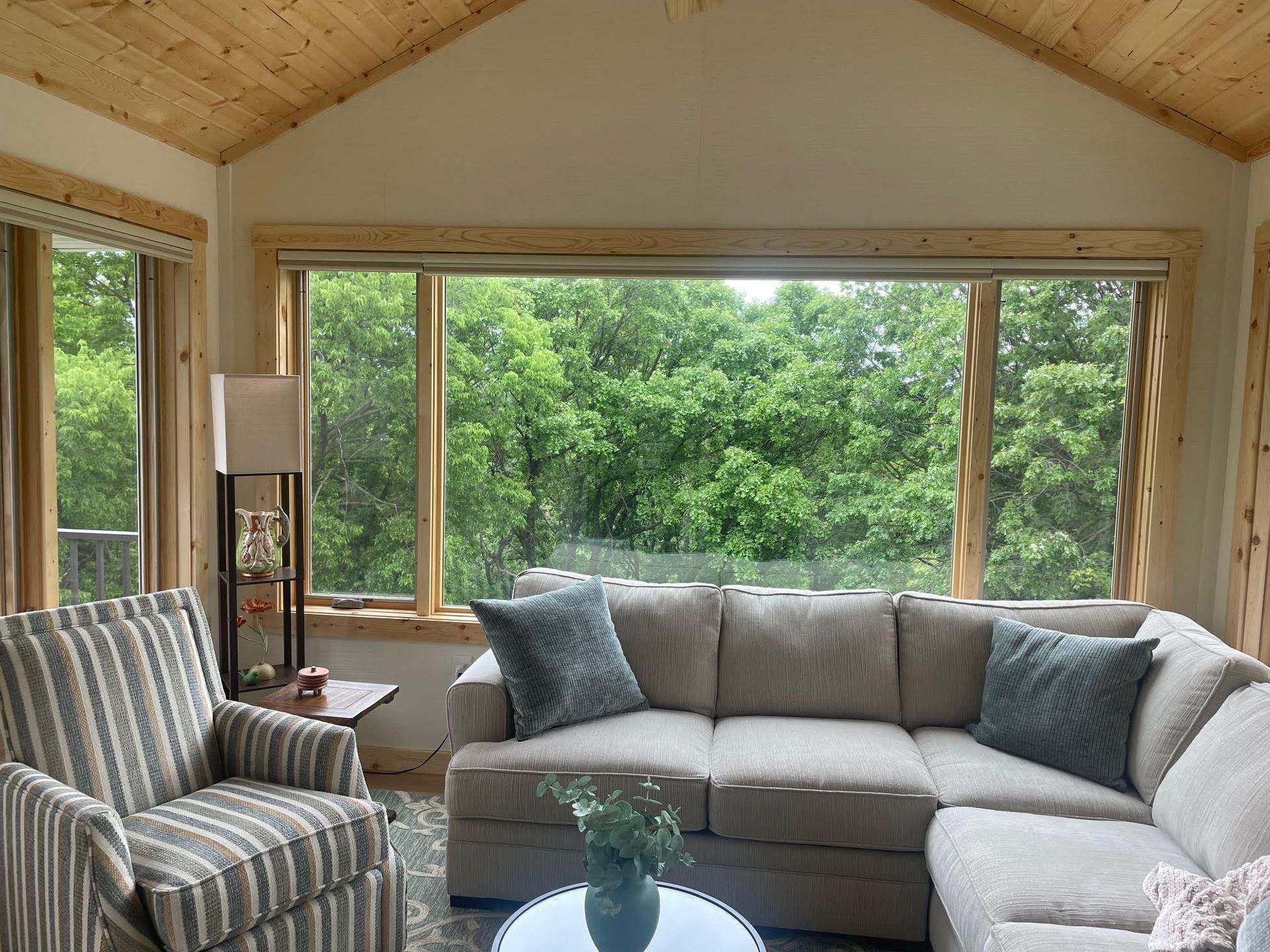 Bright sunroom with gray sectional, striped armchair, and large windows overlooking green trees