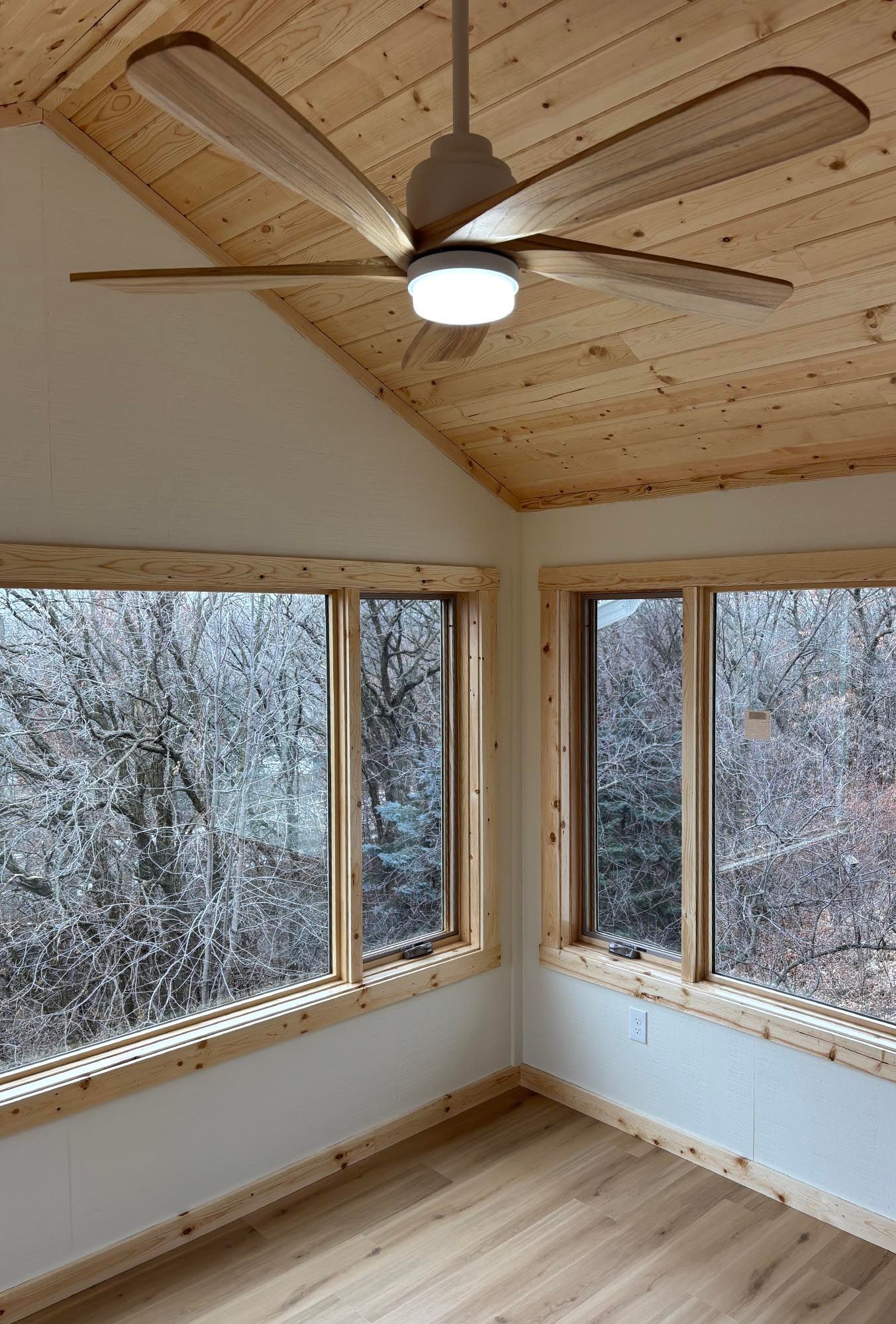 Empty sunroom with wood-paneled ceiling, ceiling fan, large windows, and snowy trees outside.