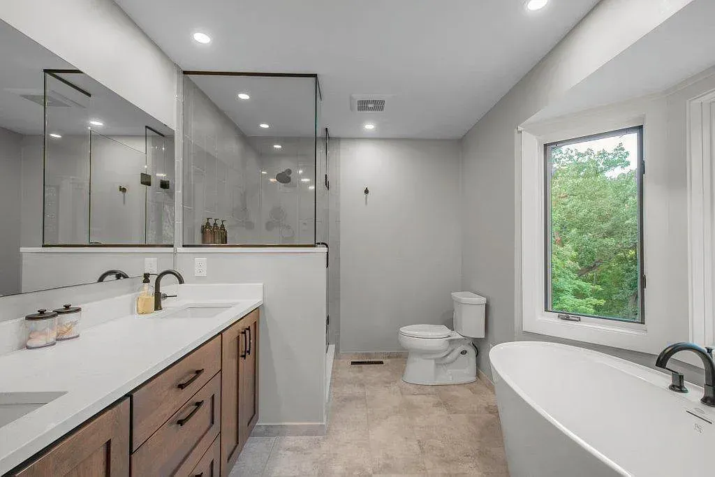 Modern white bathroom with double vanity, bathtub, toilet, and a window showing greenery.