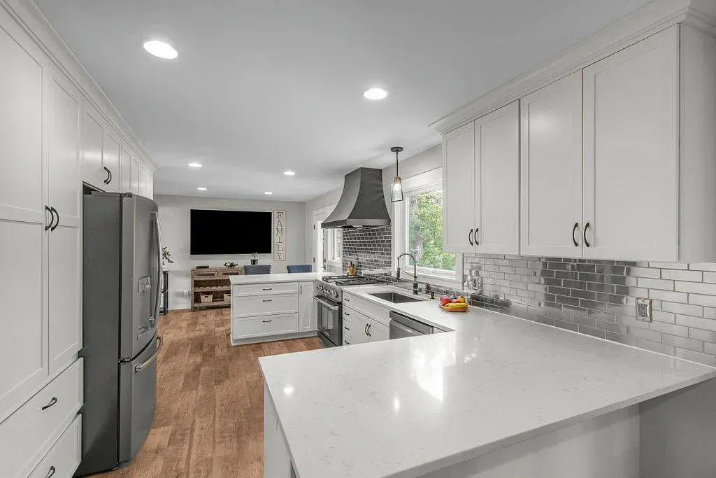 Bright white kitchen with island, stainless fridge, wood floors, and mosaic backsplash; TV visible in dining area