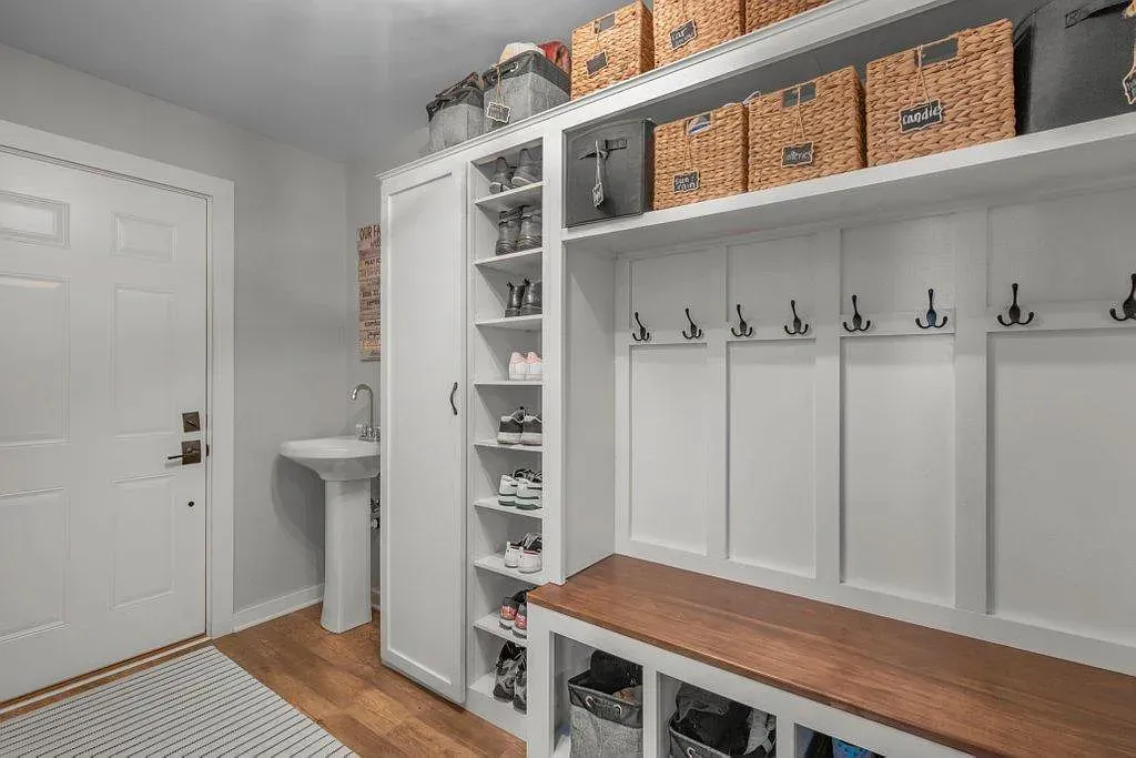 White mudroom with built-in lockers, bench, cubbies, hooks, and a small sink by the door.