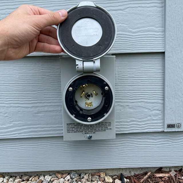 A hand holds open the metal protective cover of a gray exterior 3-prong electrical power outlet mounted on siding.