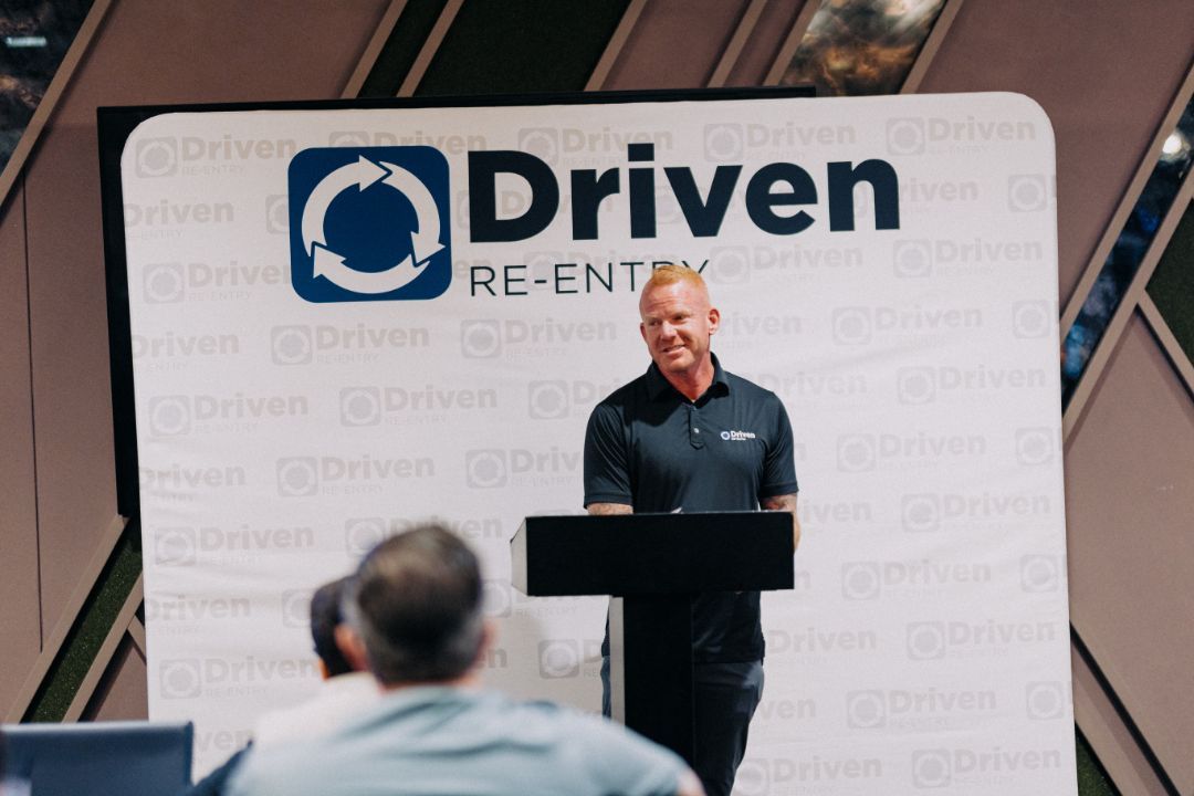 A man is standing at a podium in front of a sign that says driven.