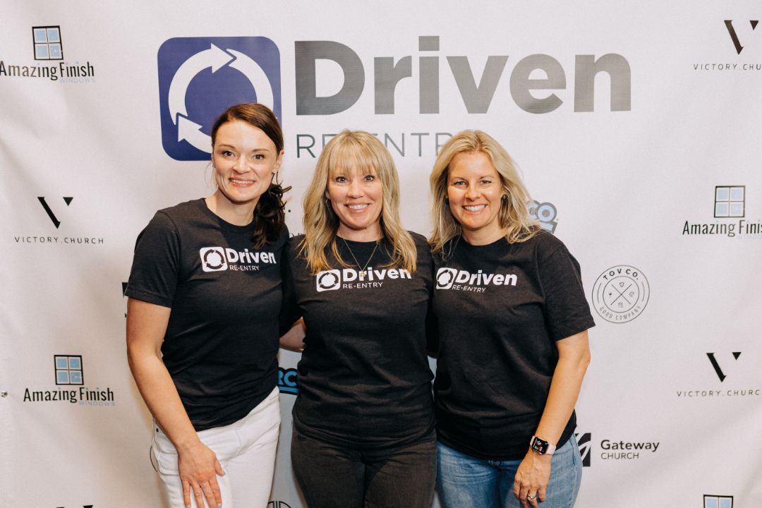 Three women are posing for a picture in front of a driven banner.