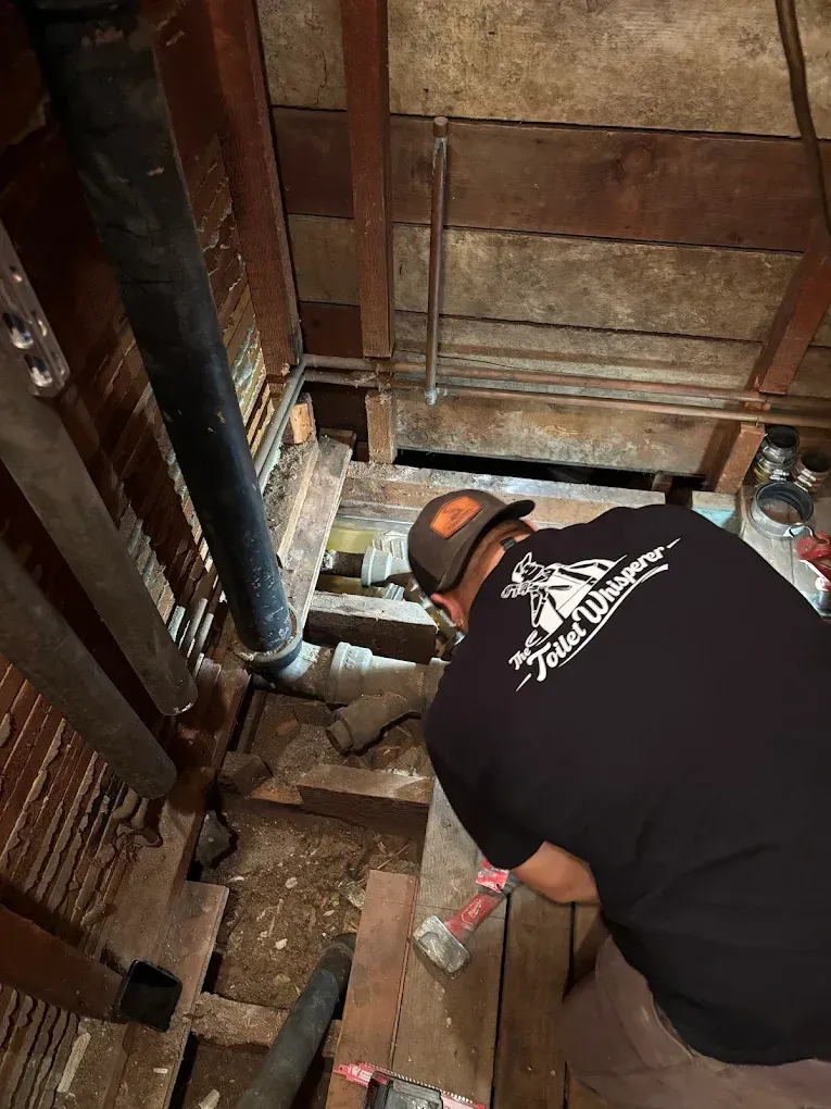 Worker repairing plumbing in a crawlspace, using tools beneath exposed floor joists.