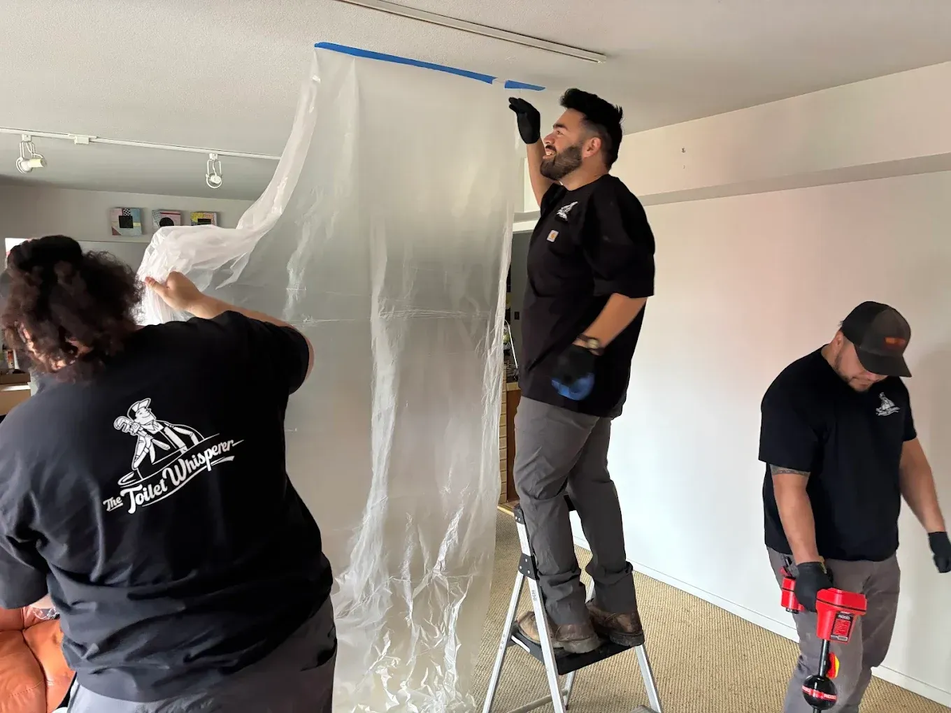 Three people painting a room, with one standing on a ladder near a plastic-covered wall.