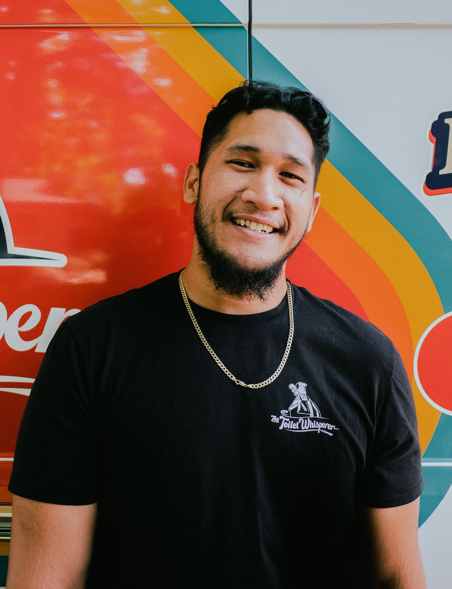 Smiling person in a black T-shirt and gold chain in front of a colorful mural.