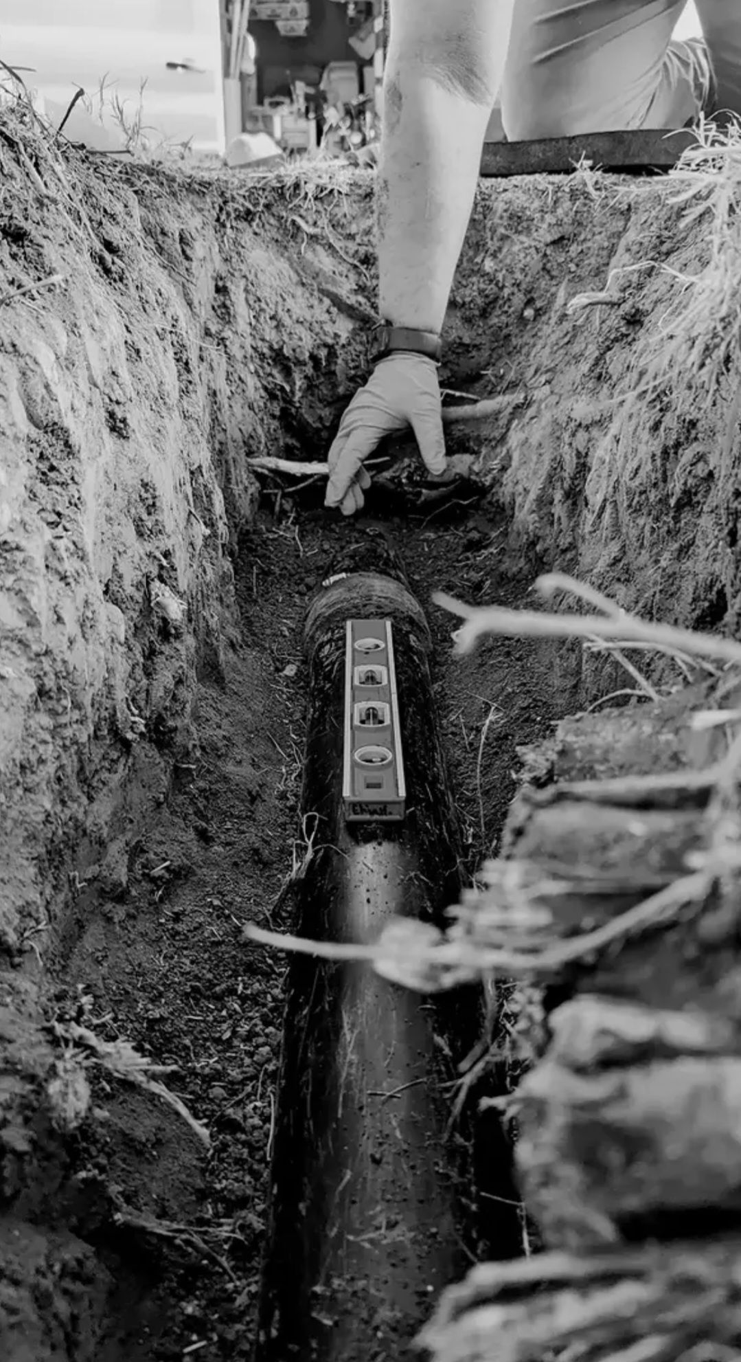 Worker in yellow hard hat and green vest standing in a dirt trench, operating a device at a construction site
