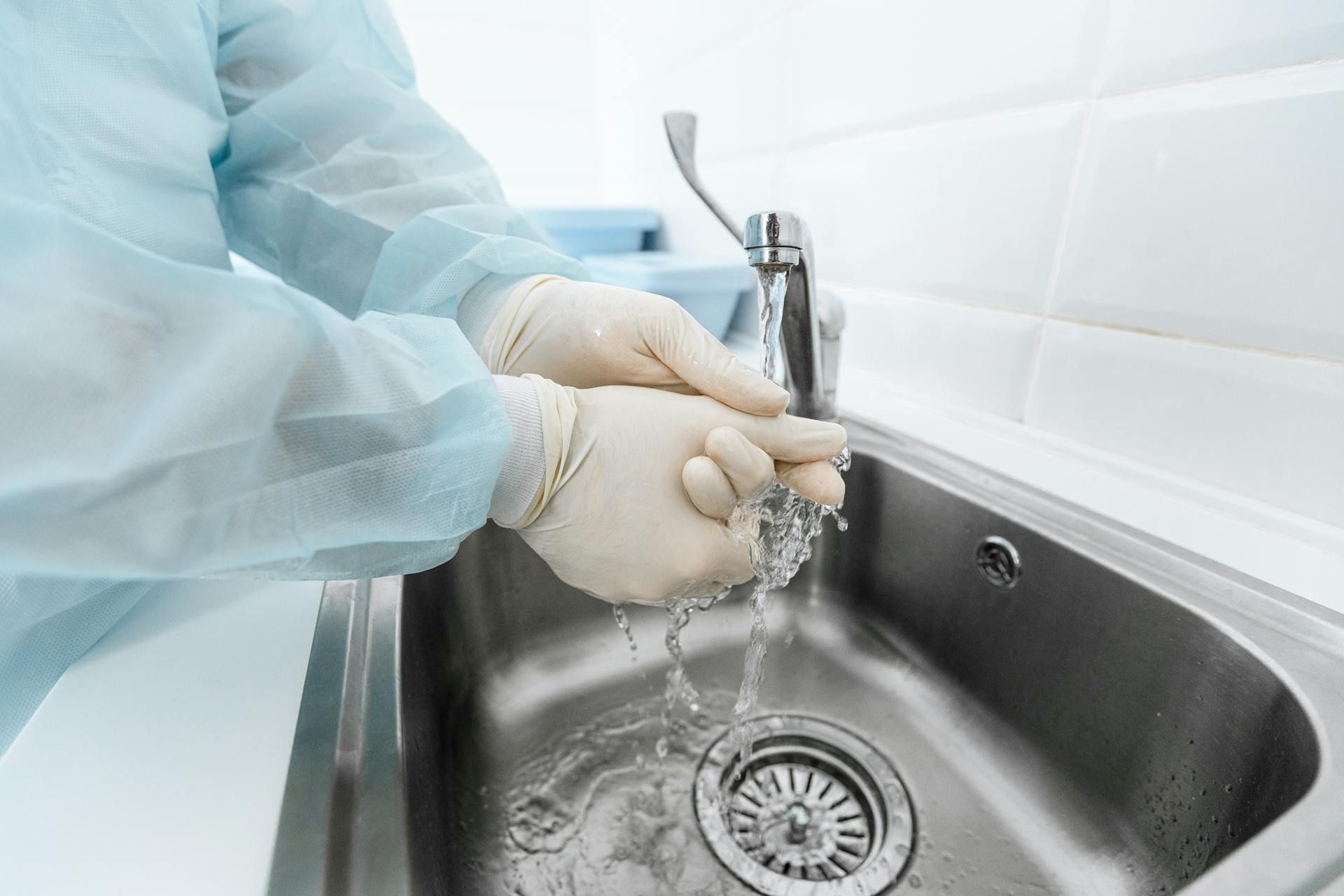 Gloved hands washing under running water in a stainless steel sink