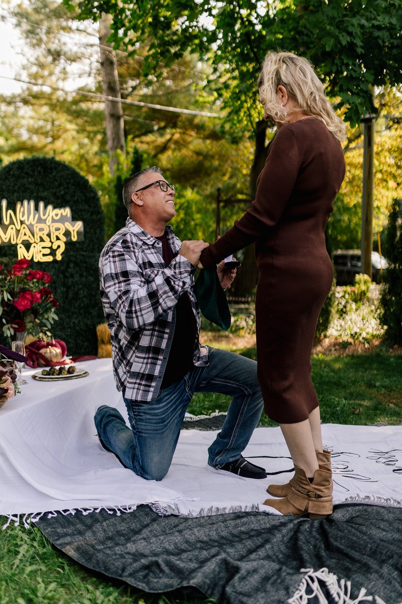 A man is kneeling down to propose to a woman in a park.
