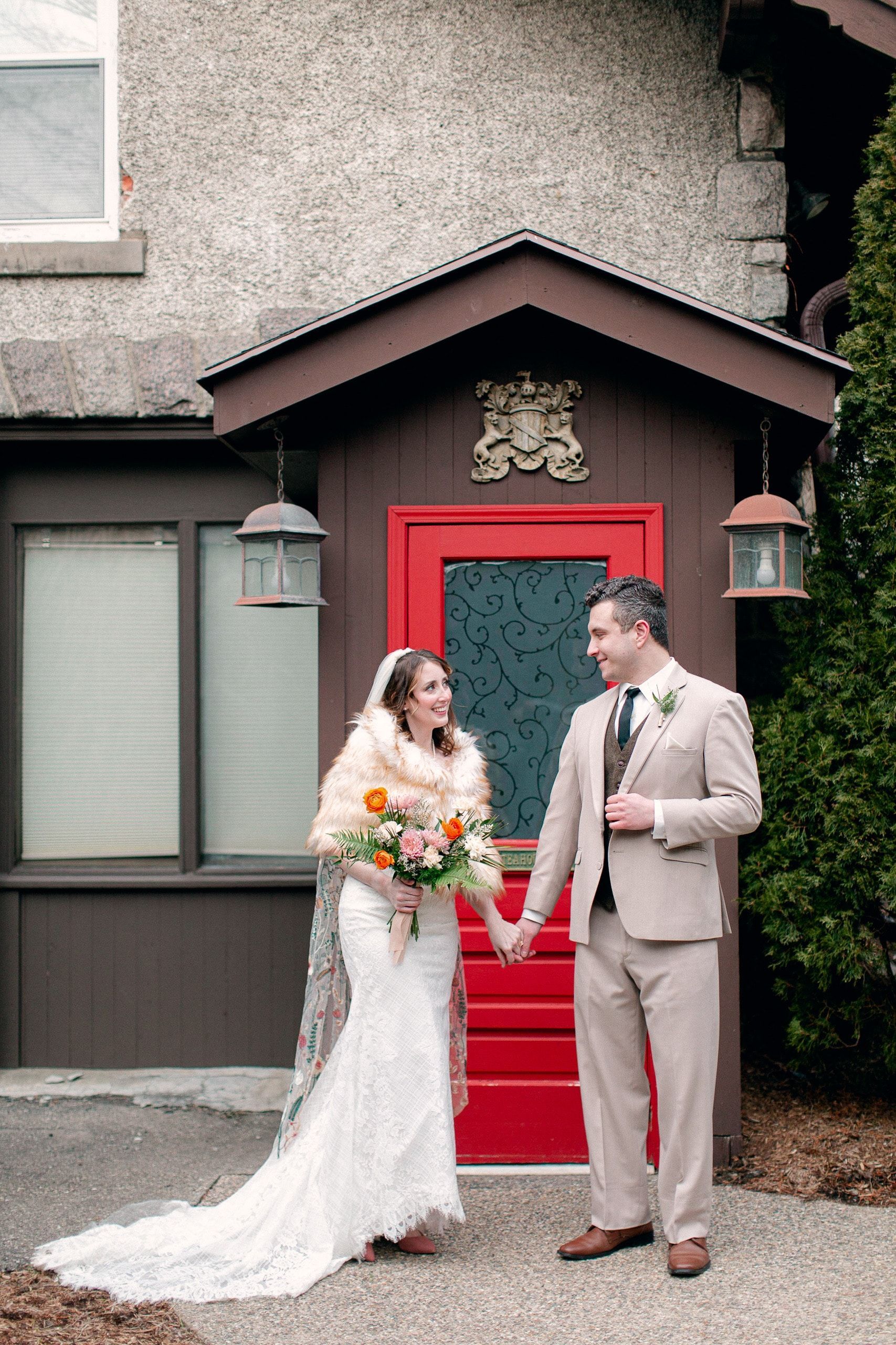 A bride and groom are standing in front of a red door holding hands.
