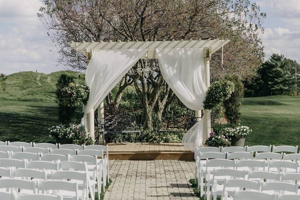 A row of white chairs are lined up in front of a pergola for a wedding ceremony.