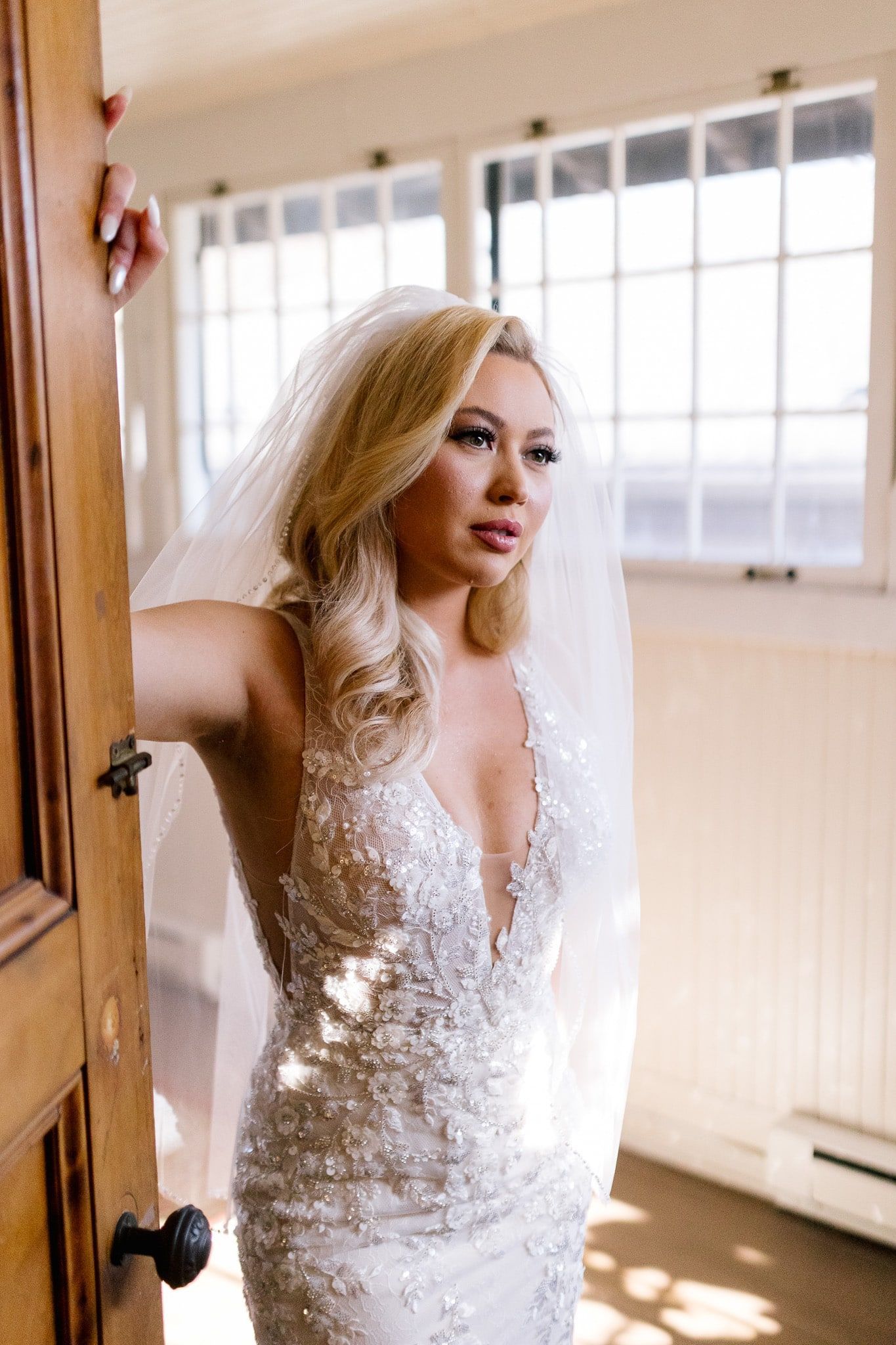 A woman in a wedding dress and veil is standing in front of a window.