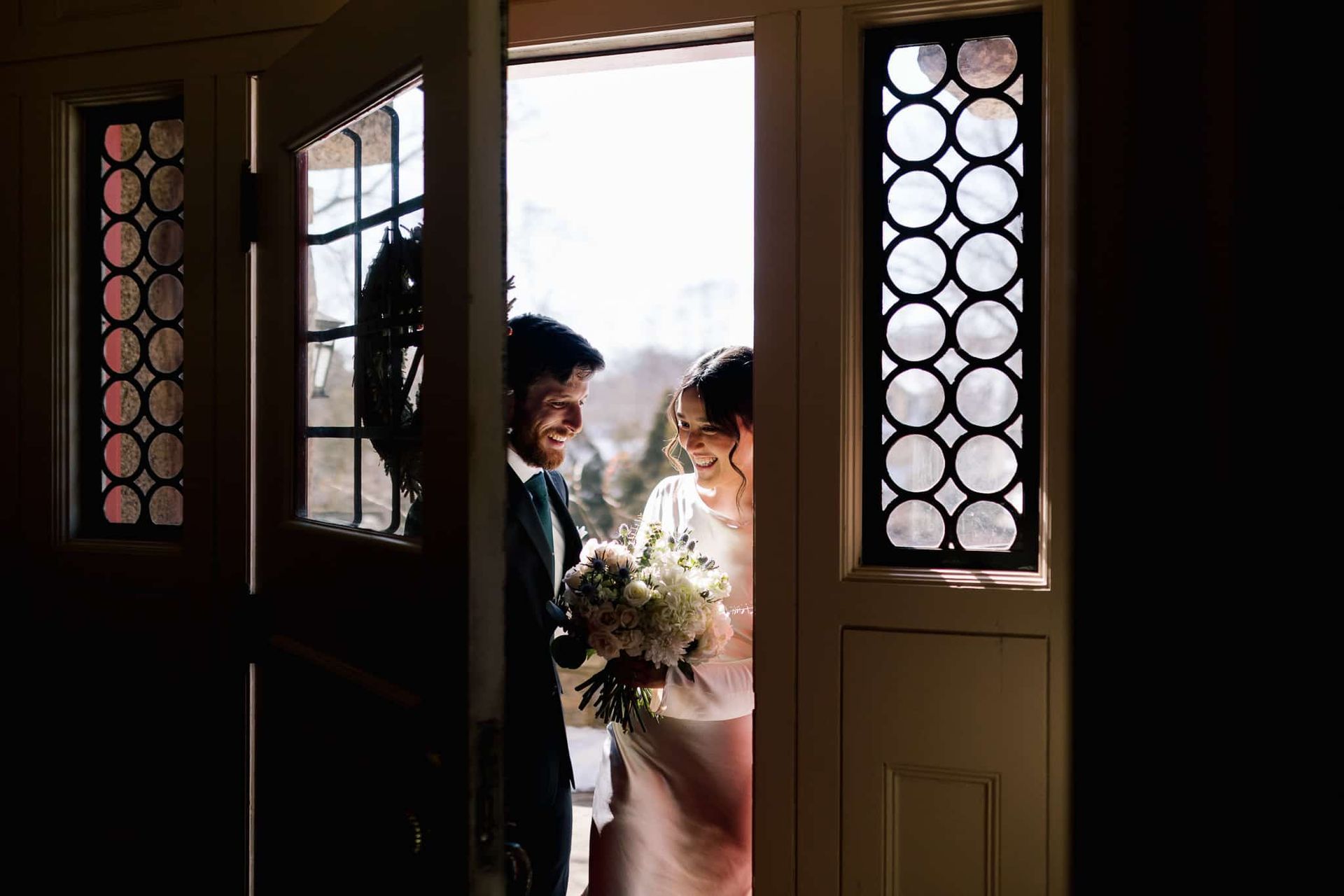 Bride and groom smile brightly, framed in a doorway, holding a bouquet, with sunlight behind them.