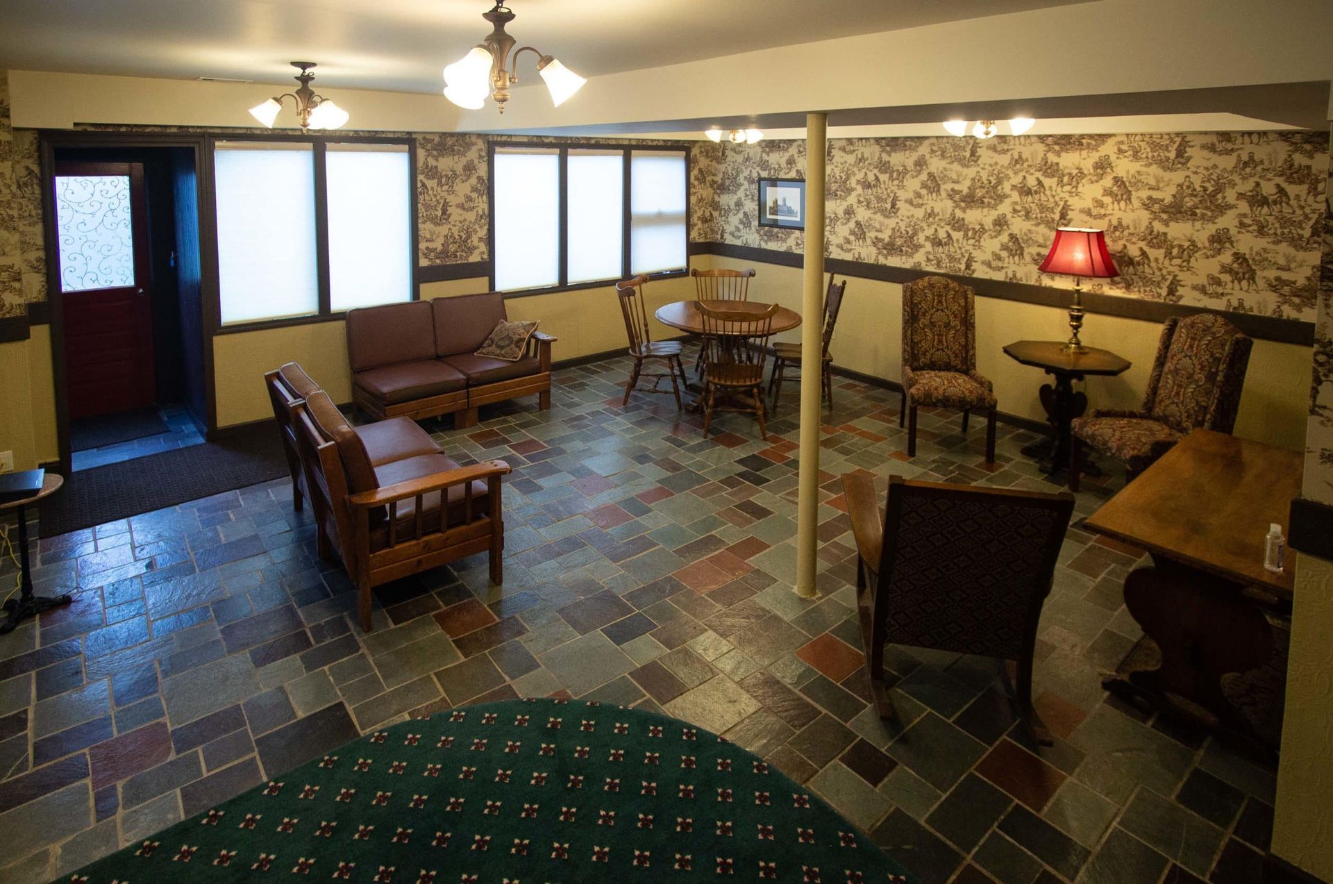 A basement lounge with various seating, round tables, and dark stone flooring. Wallpaper and windows present.