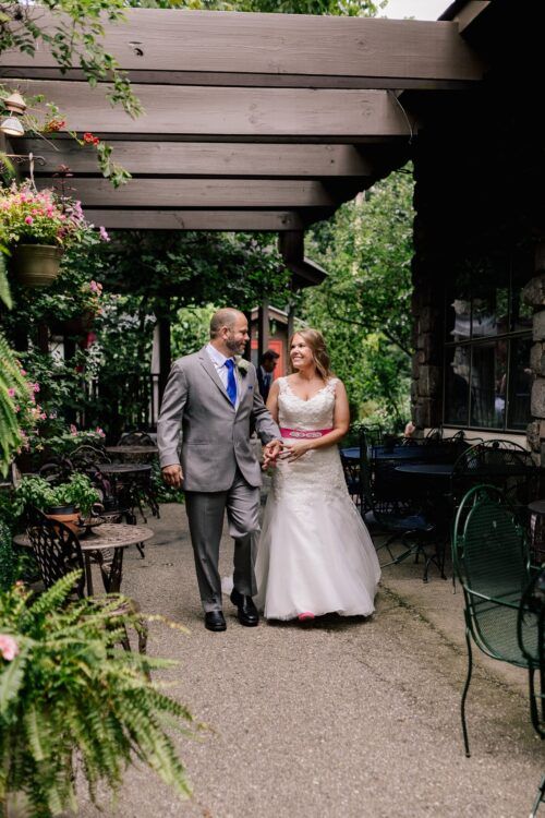 A bride and groom are walking under a pergola holding hands.
