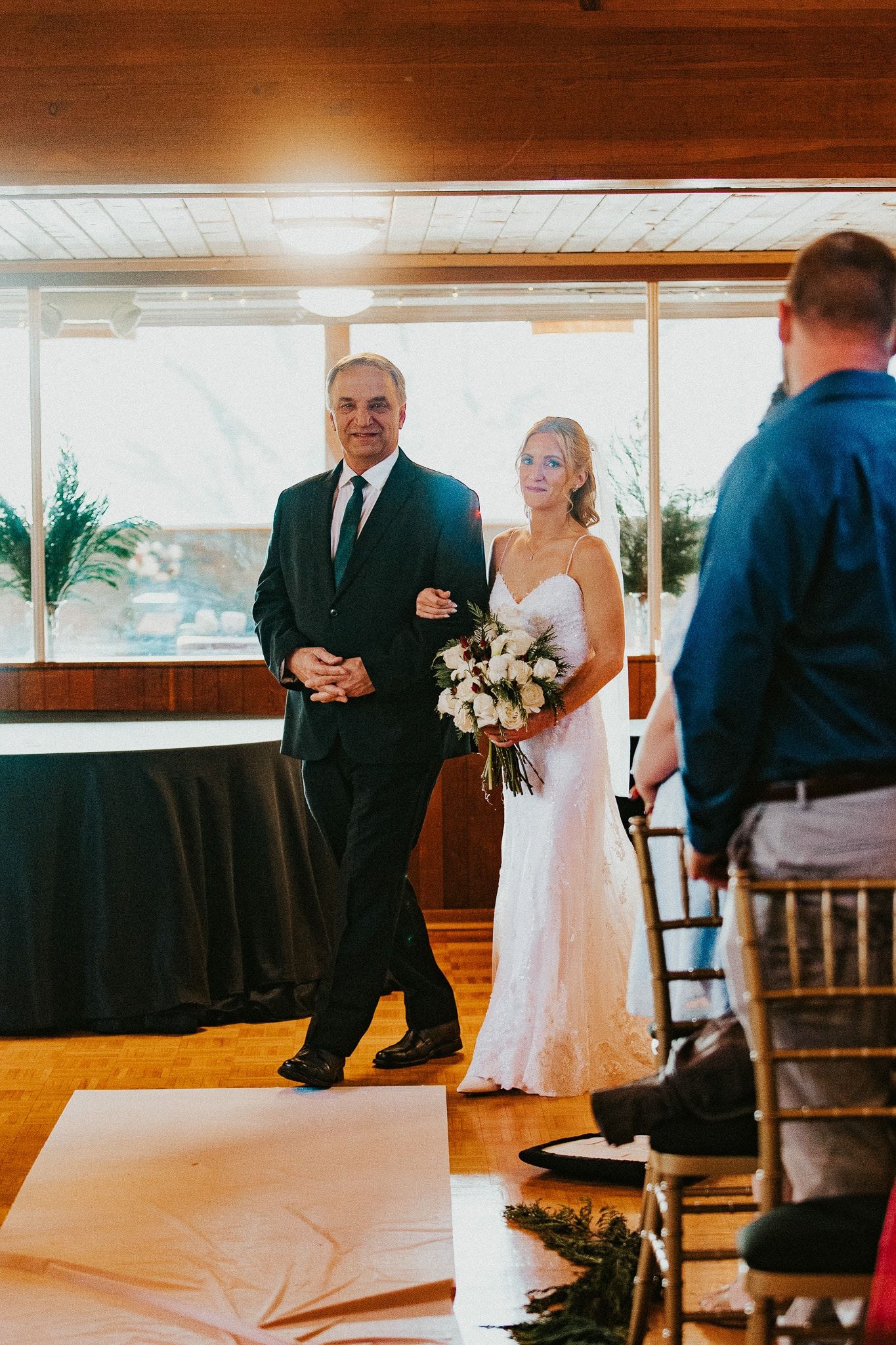 Bride and her father walking down aisle, sunlight streams in. Bride in white dress, holding bouquet. Father in suit.