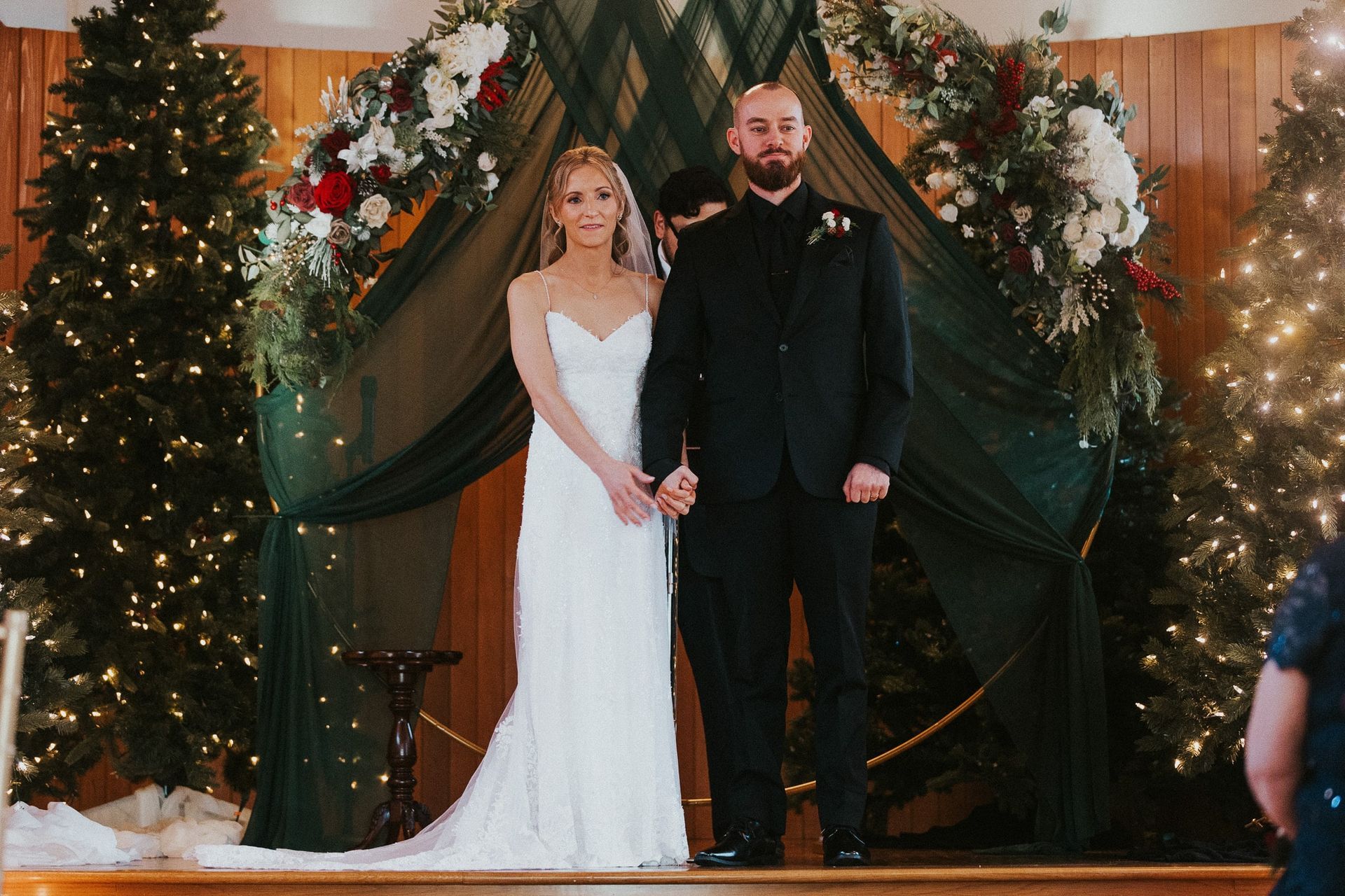 A bride and groom are holding hands during their wedding ceremony in front of christmas trees.