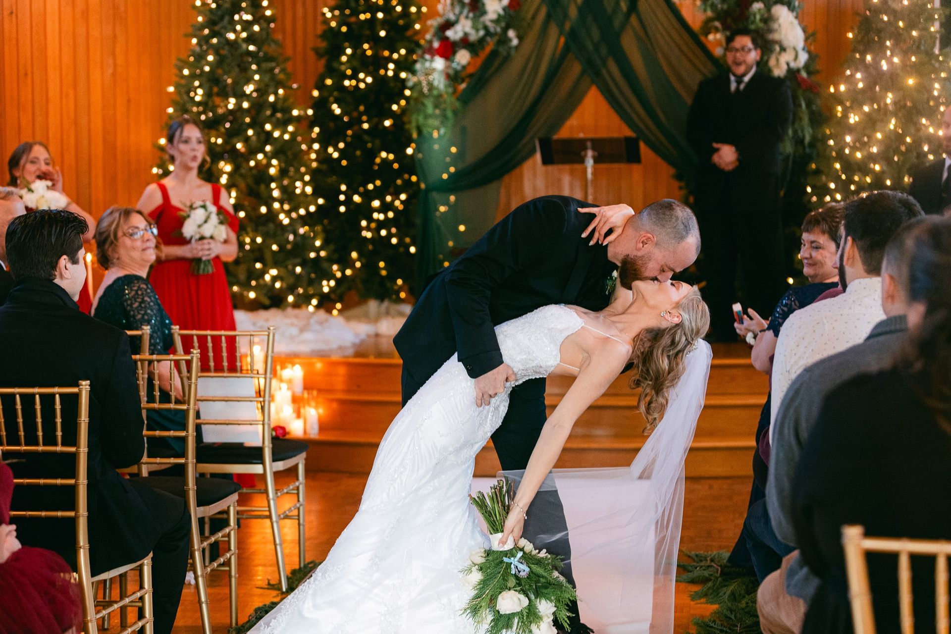 Groom kissing bride dramatically at wedding. Indoor setting with Christmas tree decor and guests.