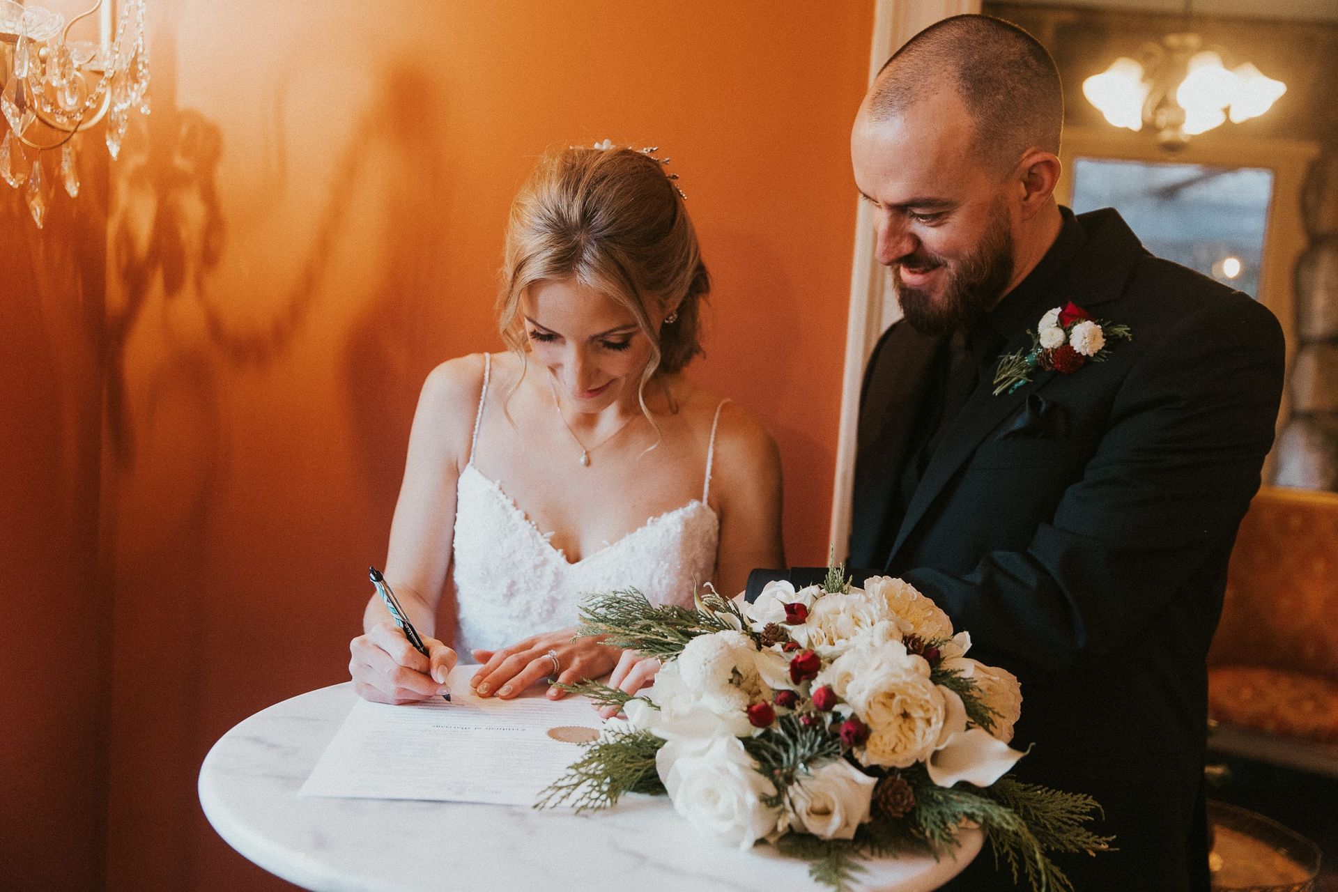 Bride signing a document with groom; a white bouquet on the table.