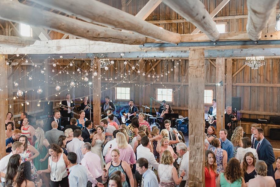 A large group of people are dancing in a barn at a wedding reception.