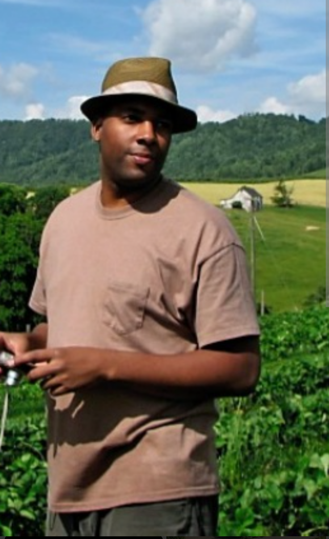 Man in tan shirt and hat standing in a green field with hills and cloudy sky in the background