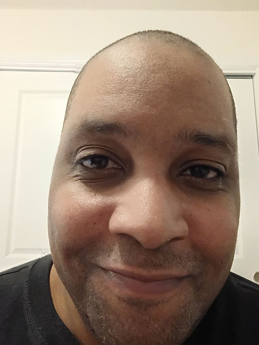 Close-up selfie of a smiling person indoors, wearing a dark shirt against a light wall.