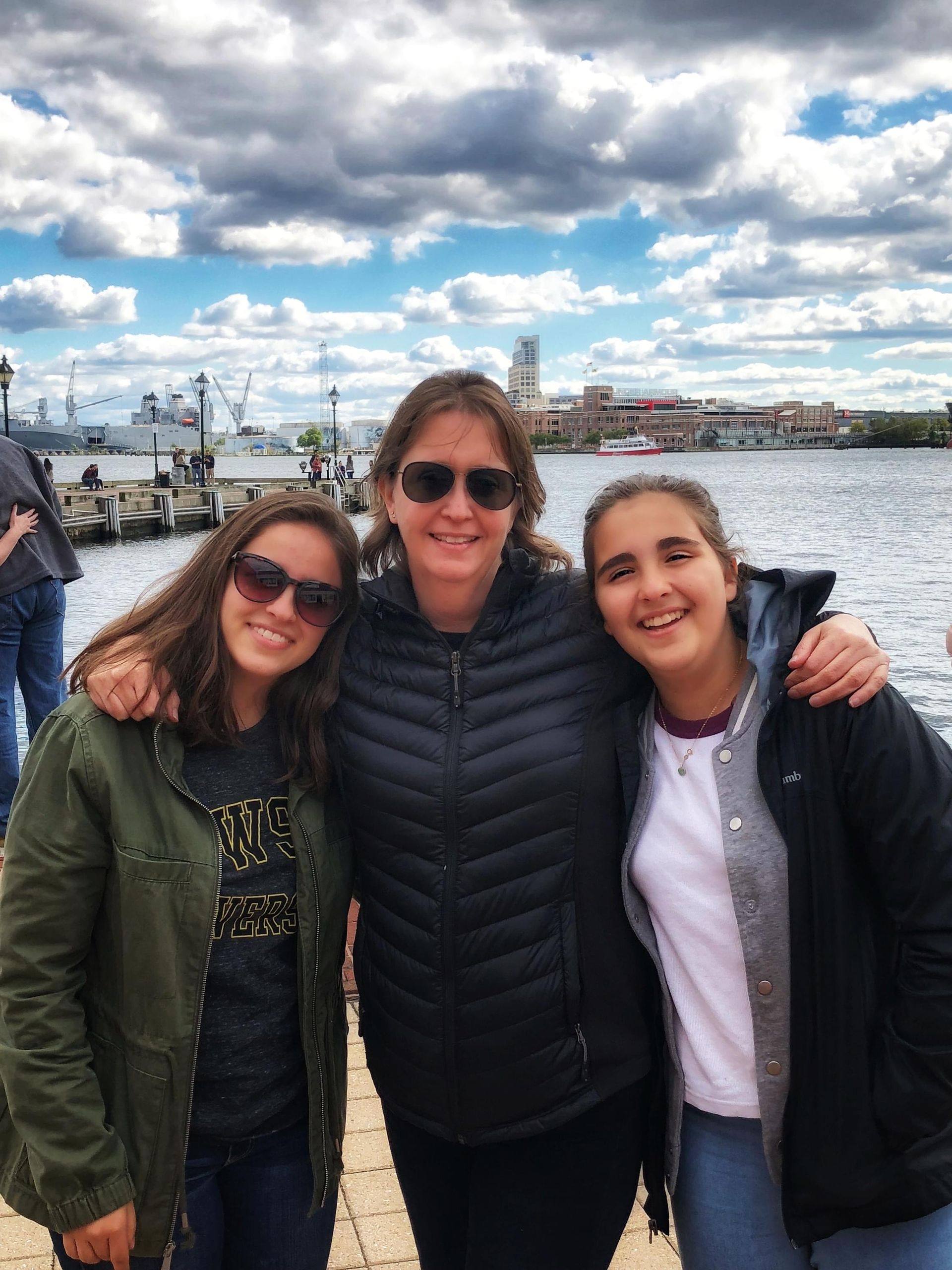 Three people smiling, posed together on a waterfront. Cloudy sky, water, and buildings in the background.