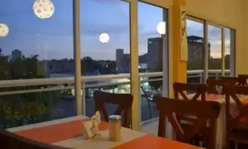 Comedor interior con mesas y sillas de madera junto a grandes ventanales con vistas a una ciudad al anochecer.