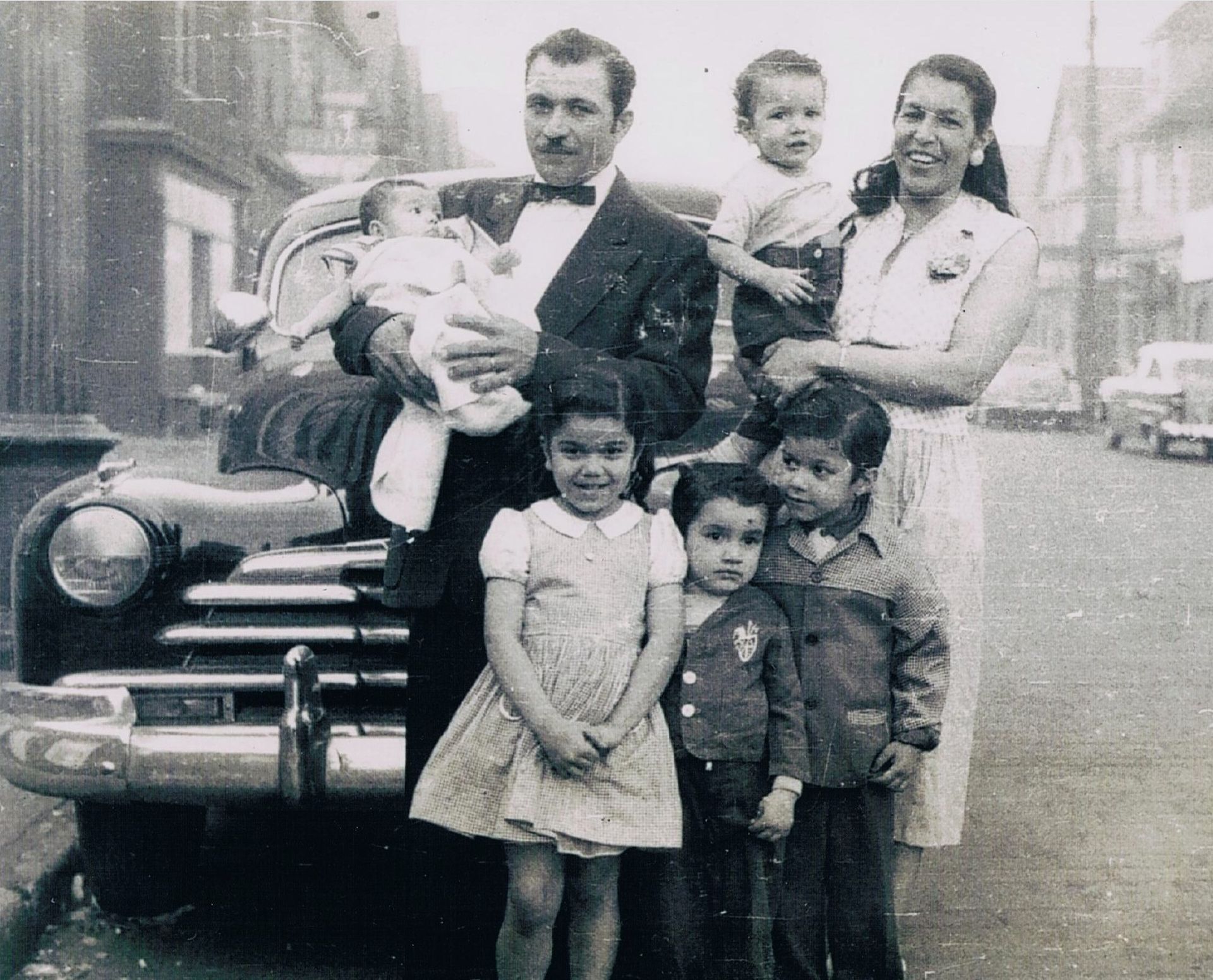 Family posing by a vintage car in a black-and-white street photo