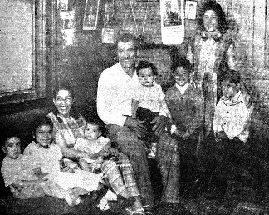 Black-and-white family portrait of seven people gathered indoors, with a seated man and children around him