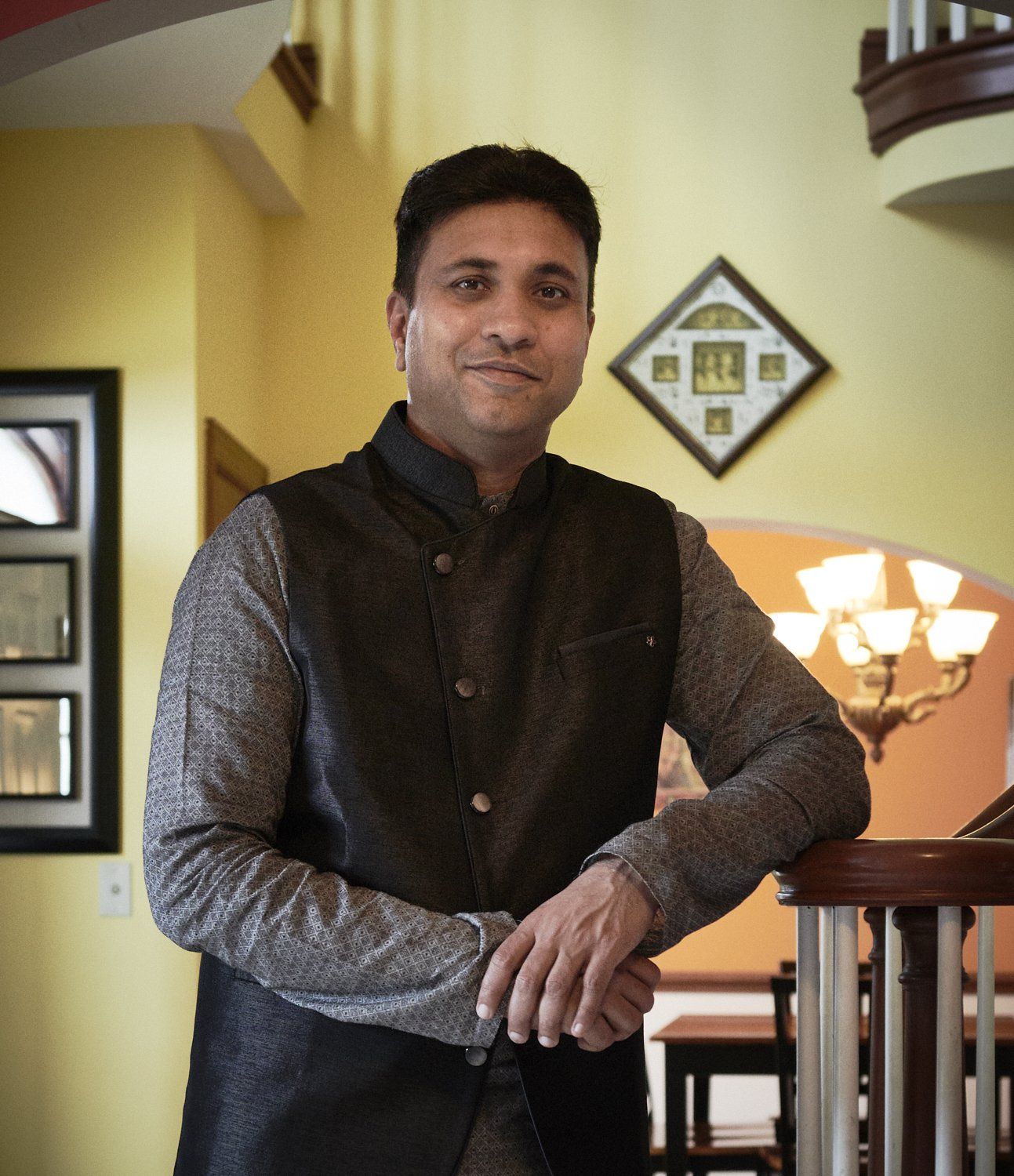 Man leaning on a stair railing in a warmly lit interior, wearing a dark vest and gray shirt