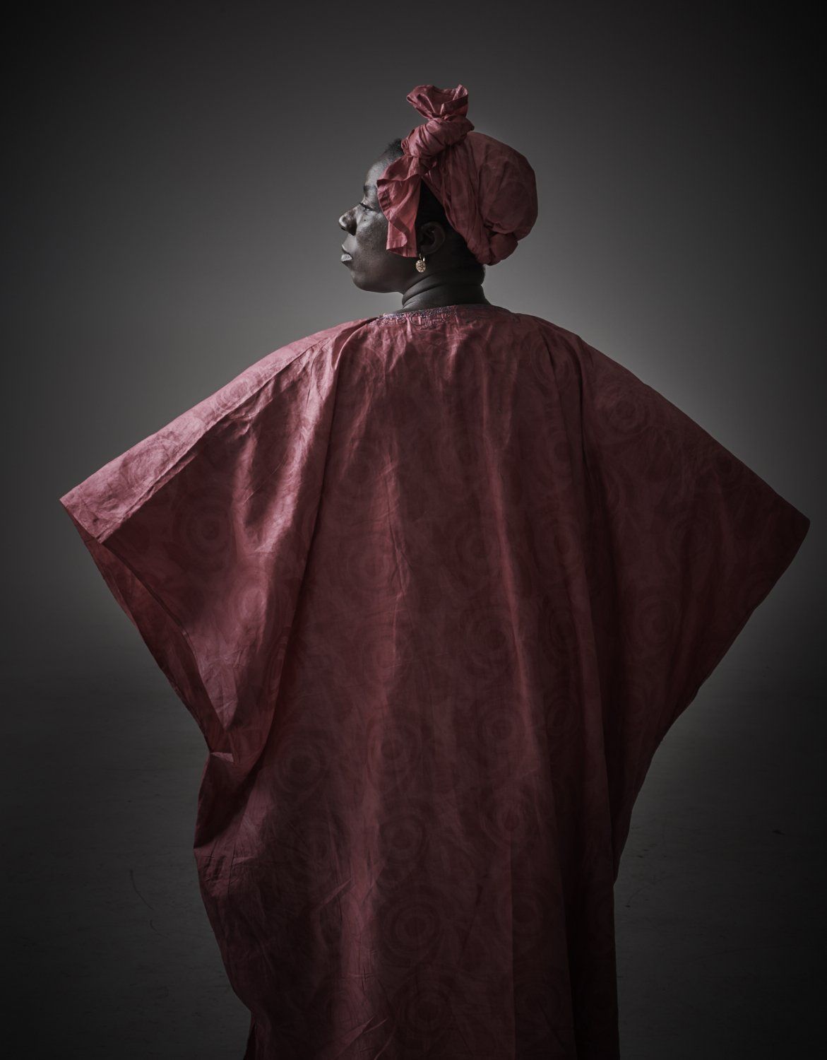 Person in a burgundy textured cape and matching headpiece, shown in profile against a dark background.