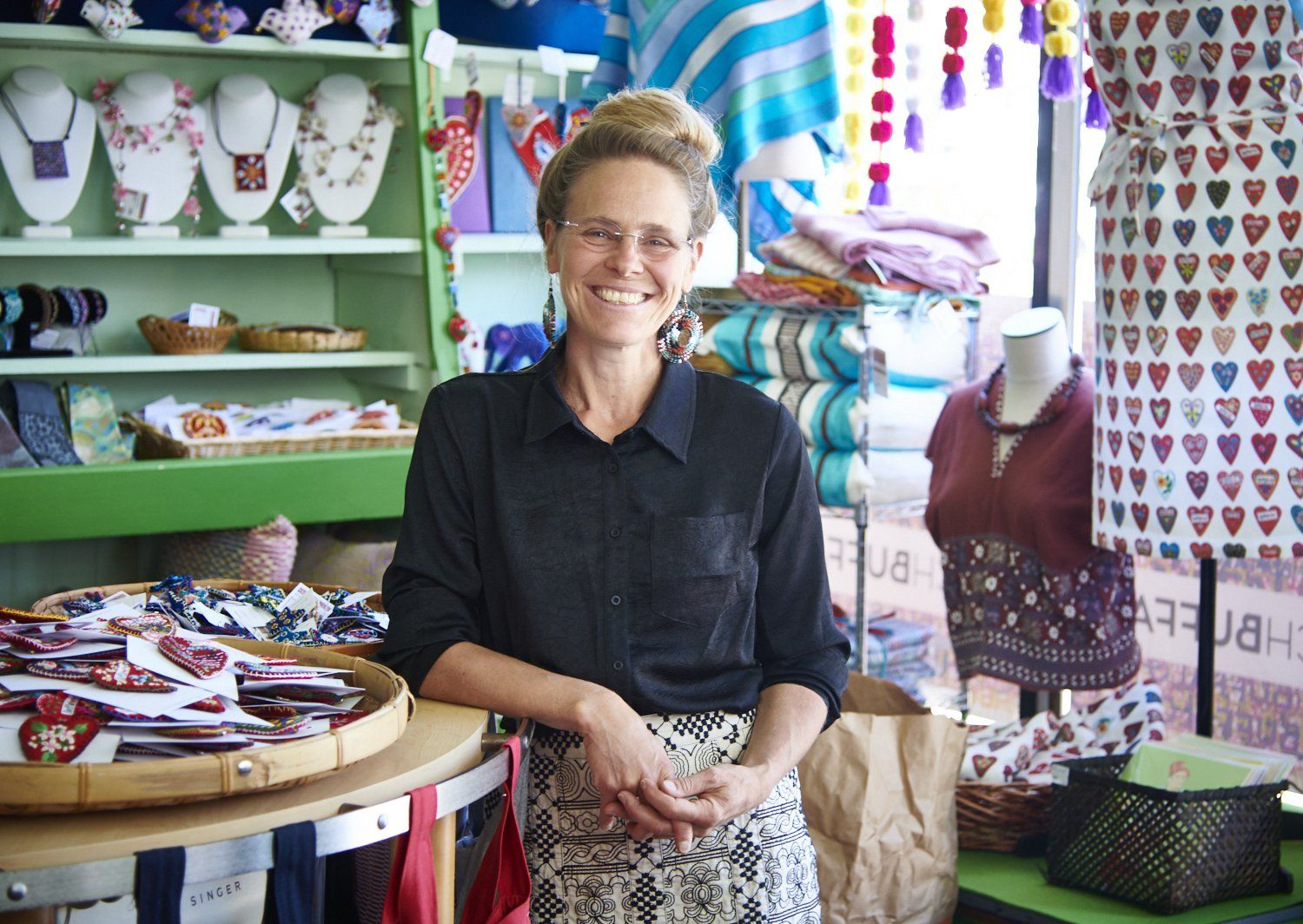 Smiling shopkeeper standing in a colorful quilt and craft shop, holding fabric by a display table.